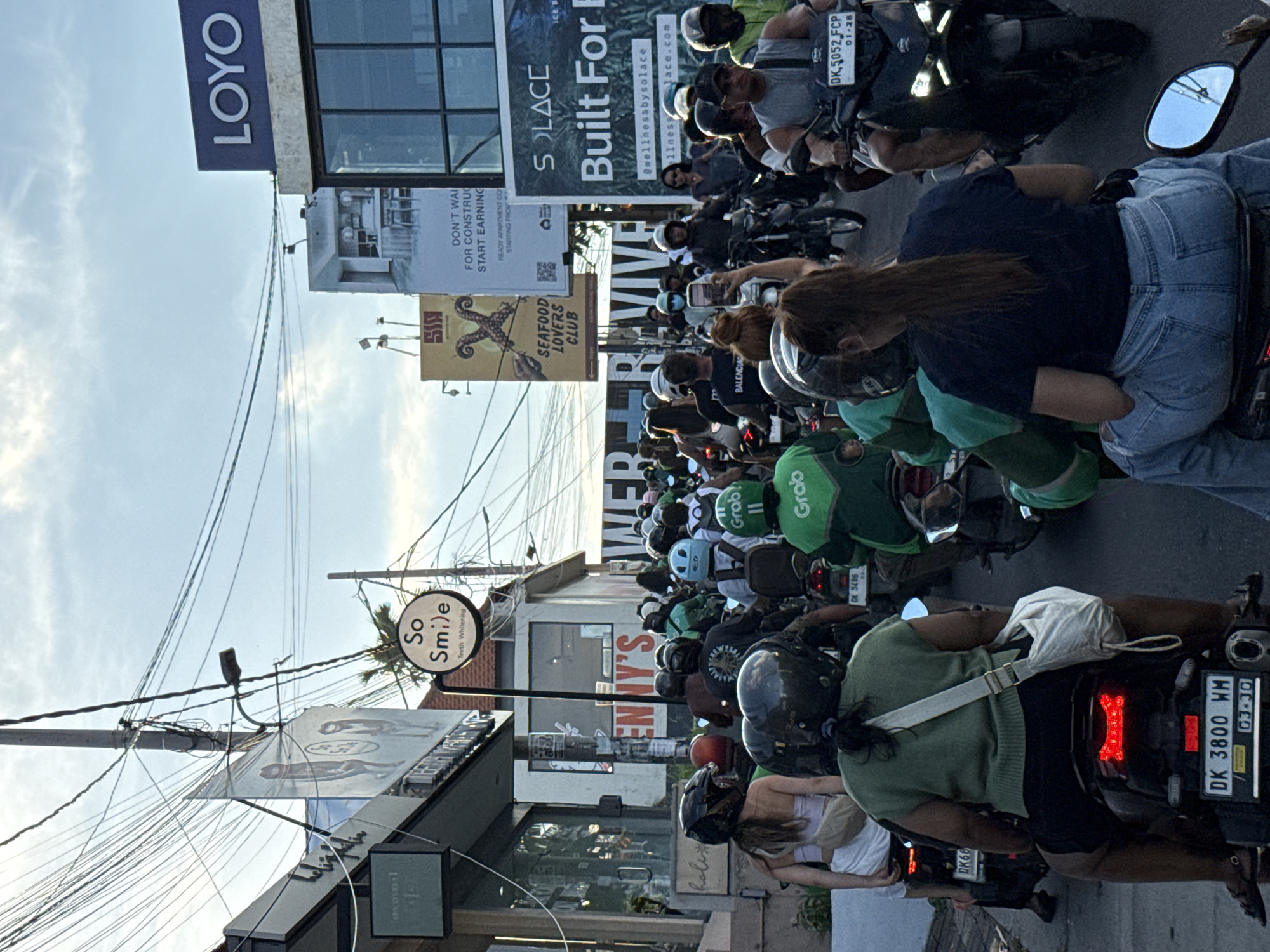 The image shows a congested street in North Kuta, Indonesia, filled with scooters and motorcycles. Many riders wear helmets, and some carry green "Grab" delivery bags. Street signs and advertisements are visible in the background, including "LOYO," "SOLACE Built For," "SEAFOOD LOVERS CLUB," "So Smile," and real estate advertisements. Power lines crisscross overhead. The scene portrays heavy traffic and the urban landscape of North Kuta.