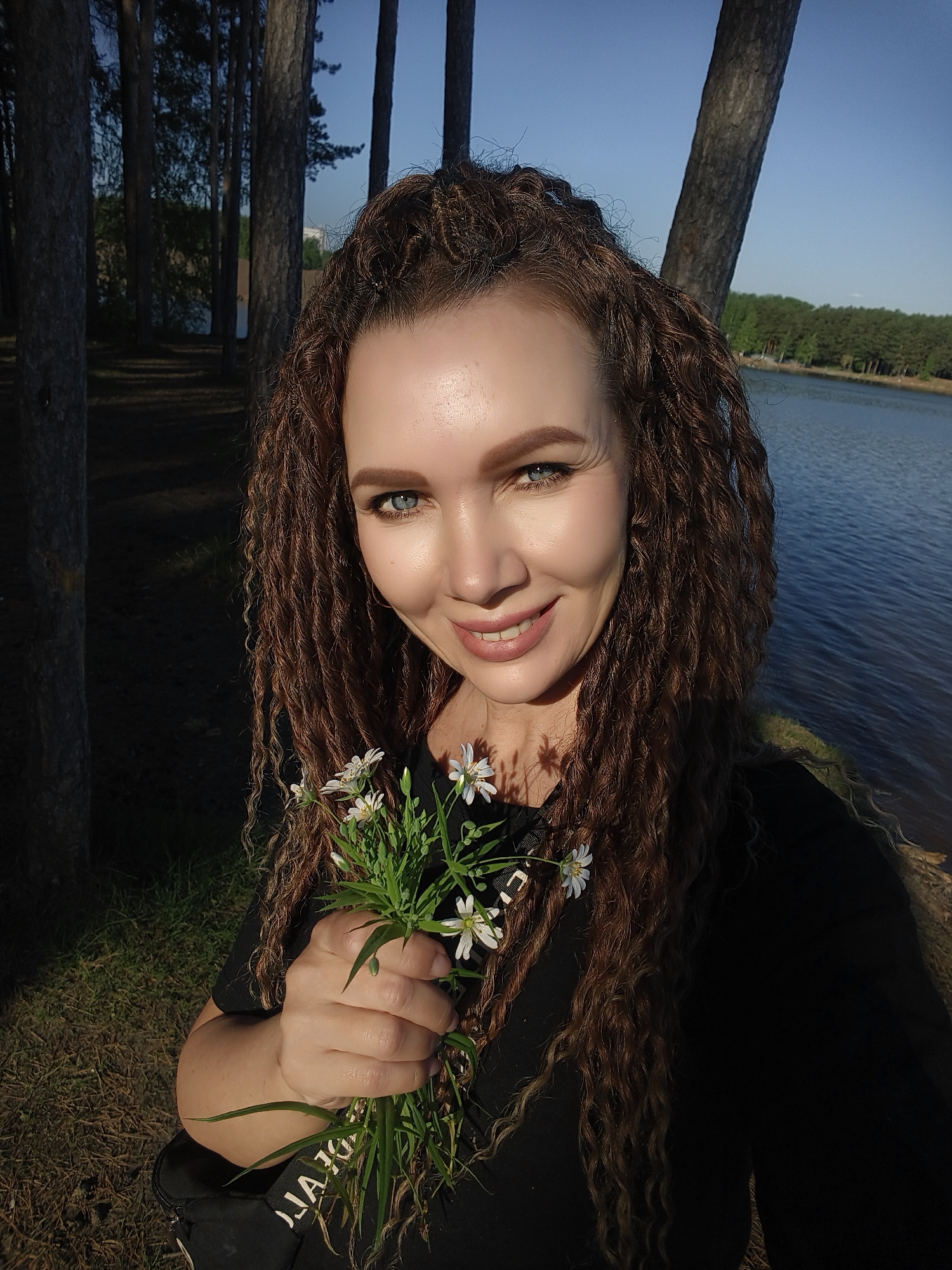 A woman with long, curly brown hair is smiling and holding a bouquet of small white flowers. She is wearing a black shirt and stands near a body of water, likely a lake, surrounded by trees. The lighting suggests it is daytime, possibly late afternoon. The background includes a grassy area and a treeline, creating a natural setting. Unknown, Unknown.