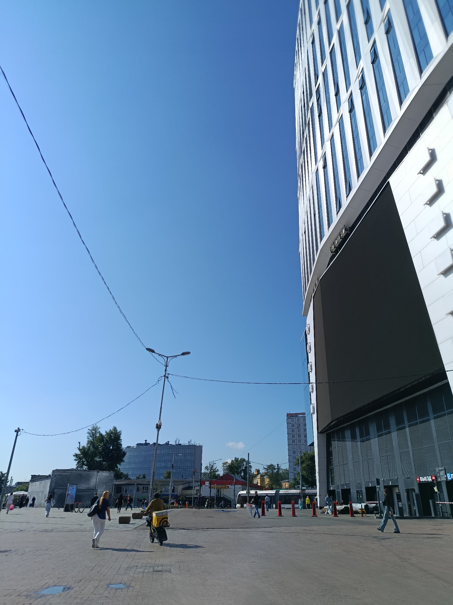 In Moscow, Russia, a bright, clear day is depicted with a wide, paved plaza in the foreground. A modern, tall, white building with many blue-tinted windows dominates the right side of the frame, featuring a large, dark digital display screen. To the left of this building, a lamppost with two lights stands tall, with electrical wires stretching across the sky. In the midground, several people are walking on the plaza. One person is riding a scooter with a yellow delivery box on the back. Further back, a row of buildings, including a glass-fronted structure, are visible, along with trees and a tram. The overall scene suggests a bustling urban environment under a cloudless blue sky.