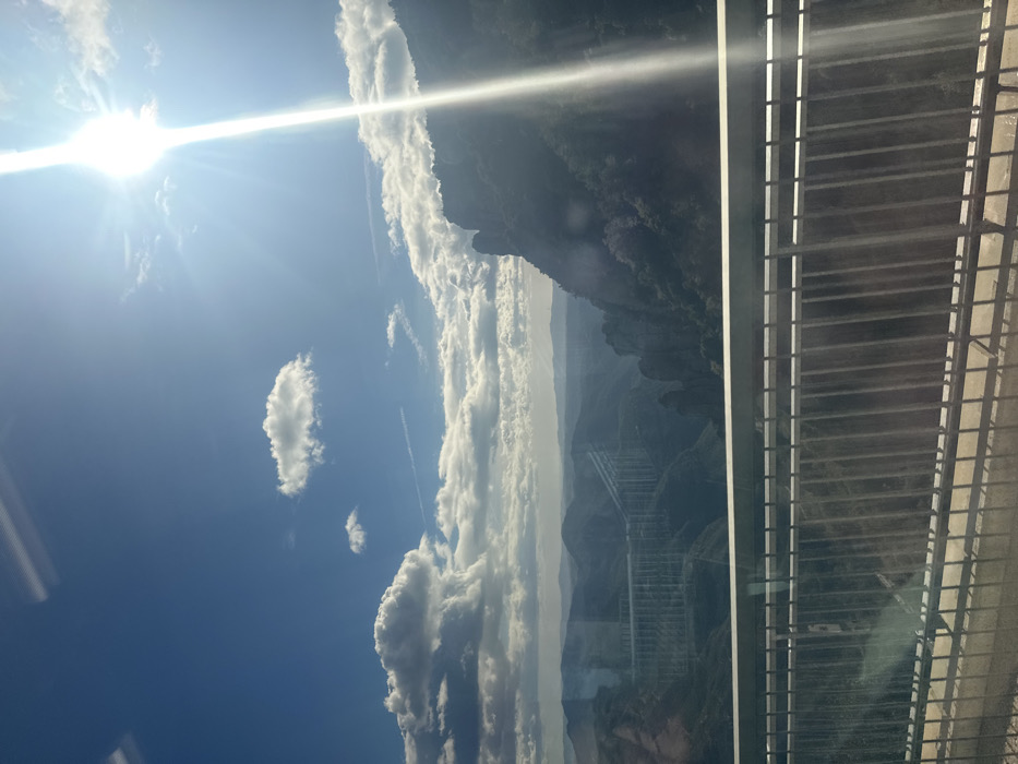 The image displays a panoramic view of the Vacarisses Viaduct (Viaducte de Vacarisses) in Vacarisses, Spain, under a bright, partly cloudy sky. The scene is observed from an elevated position, likely from a vehicle, indicated by a metal guardrail in the lower foreground. The sun, located in the upper right, emits a strong vertical flare across the image, casting intense light on the mountainous landscape.
In the midground, a deep valley is flanked by rugged, forested mountains, with discernible rock formations on the right. Spanning the valley on the left, the Vacarisses Viaduct is prominently visible, characterized by its multiple concrete arches or pillars supporting a broad roadway. The viaduct appears lighter in tone against the darker, shaded mountainsides. Scattered cumulus clouds and faint contrails are present in the blue sky. Distant mountain ranges form the hazy horizon. No individuals, specific actions, or interactions are observable.