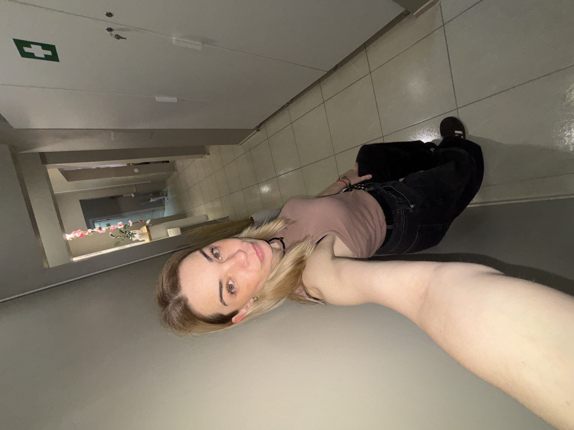 A waist-up view of a woman taking a selfie, facing the camera while standing in a hallway. The woman is wearing a pink tank top, black jeans, and a black choker necklace. She has fair skin, blonde hair, and hazel eyes. A medical kit sign is seen on the ceiling. A floral arrangement can be seen at the end of the hallway. The location is unknown.