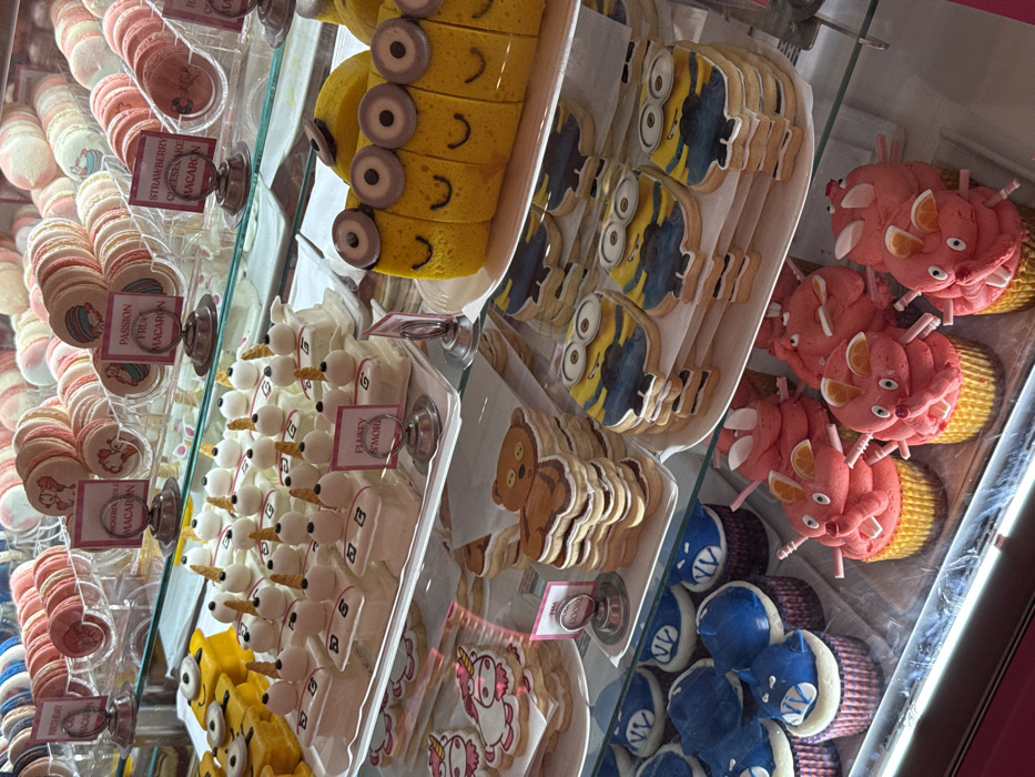 This image depicts the interior of a brightly lit display case within a dessert shop in Orlando, United States of America, showcasing an array of confectionery items across four glass shelves.
On the top shelf, multiple varieties of macarons are presented in clear acrylic cylindrical containers. Flavors visible on signs include "BIRTHDAY MACARON," "MOCHI MOLI MACARON," "PASSION FRUIT MACARON," and "STRAWBERRY CHEESECAKE MACARON." The macarons are pastel-colored, predominantly pinks and whites.
The second shelf features two distinct dessert types. To the left, a long, segmented yellow cake or log-roll is decorated to resemble a series of Minion characters, complete with brown goggle-like eyes and drawn-on smiles. To the right, a tray holds approximately 20 individual square desserts, each topped with a toasted marshmallow and a small golden cone, labeled "FLUFFY S'MORE." These appear to be s'mores bars.
The third shelf displays stacks of themed sugar cookies. On the left, a stack of unicorn-shaped cookies with white, pink, and gold frosting, and adjacent to them, a stack of teddy bear-shaped cookies with brown and white frosting. On the right, stacks of Minion-shaped sugar cookies, decorated in yellow, blue, and black.
The bottom shelf contains two types of cupcakes in patterned wrappers. On the left, several cupcakes with blue frosting sculpted to resemble monster heads or faces, some with white accents suggestive of eyes or fangs. On the right, multiple cupcakes with pink frosting sculpted into pig or mouse-like forms, featuring pink ears and snouts, with some adorned with small orange fruit slices.
The entire scene is illuminated by internal lighting, highlighting the confections. No individuals are visible in the image.