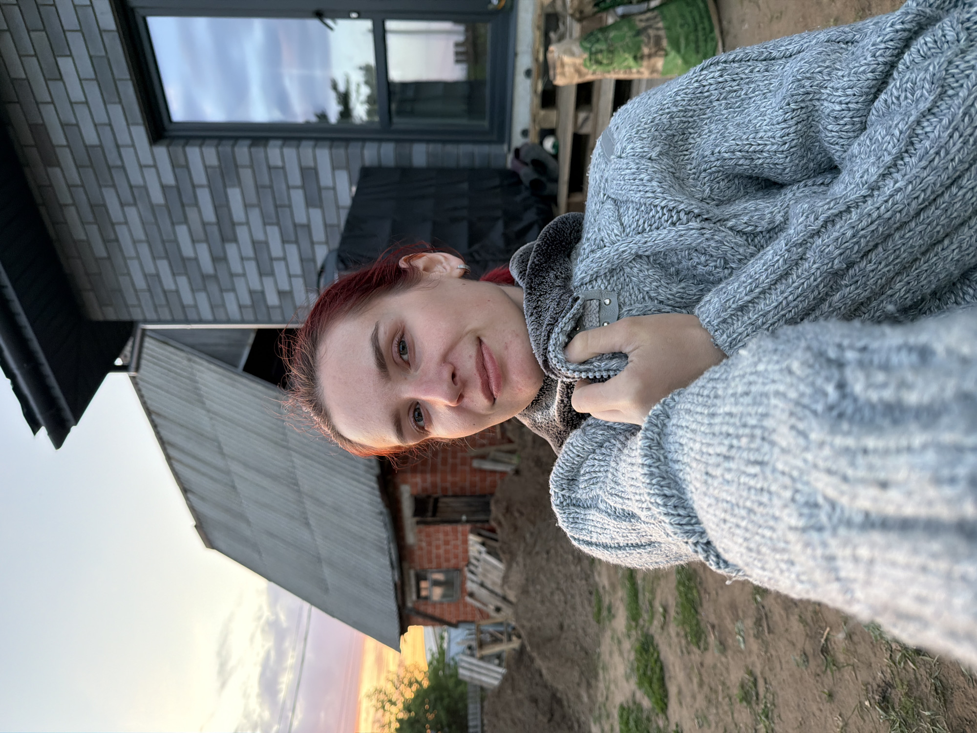 A medium shot of a fair-skinned person with reddish hair wearing a gray knitted sweater. The person is facing the camera and slightly tilted. In the background is a combination of modern and older structures. To the left is a building with gray brick, a gray door, and a modern, dark-framed window reflecting the sky. Further to the left, there is a sloped gray roof. Behind the person's head and to the left is a partially visible building with brick walls and a wooden structure next to it. The ground is dirt with some sparse grass and greenery. A sunset is visible near the bottom of the frame. This scene takes place in Chaikovsky, Russia.