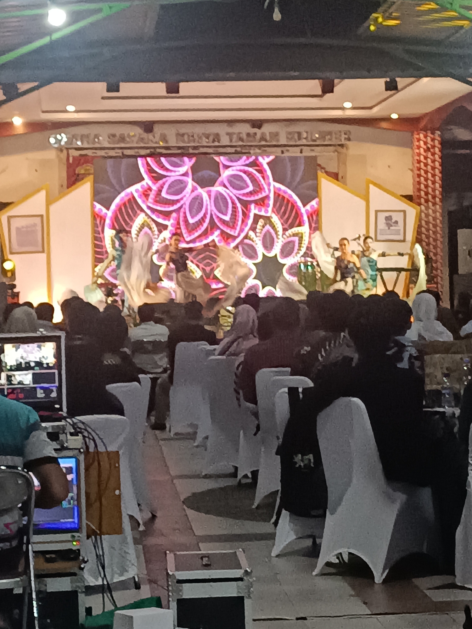 The image depicts an indoor event space in Condongcatur, Indonesia, likely a community hall or auditorium, with a stage at the rear. The stage features a large LED screen displaying a vibrant pink and purple floral pattern. Several performers, appearing to be dancers and possibly musicians, are on the stage. Some are in motion, with flowing fabric, suggesting a dance performance. Two individuals are positioned near a keyboard-like instrument. In the foreground, an audience is seated in rows of white-covered chairs. A person in a teal shirt is visible to the left, facing a video control setup with multiple screens and cables. Equipment cases are also present in the lower part of the image. The overall atmosphere suggests a cultural or celebratory gathering.
