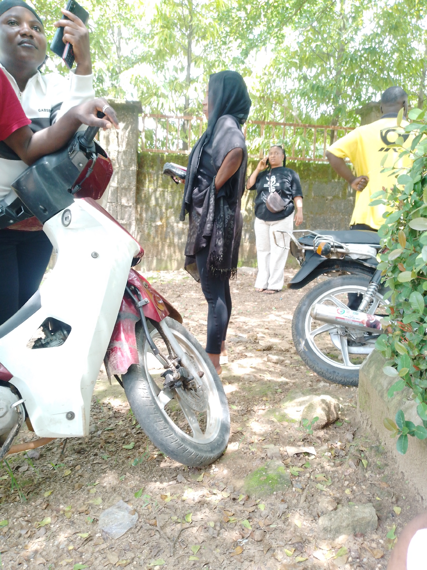 The media file shows a group of people standing outdoors, near parked motorcycles. In the foreground, a white and red motorcycle is partially visible, with its front wheel and fender in focus. To the left, a person wearing a red shirt and a white jacket with "GABSE" printed on it is seated on the motorcycle, holding a dark-colored phone.
Further back, another person is standing, wearing a black hijab and a dark-colored scarf. To their right, a woman in a black t-shirt and white striped pants is on the phone, with a small brown bag slung across her chest. Behind her, a person in a bright yellow t-shirt stands facing away from the camera.
The setting appears to be outdoors, with trees and foliage in the background, suggesting a natural or semi-rural environment. The ground is covered in dirt and fallen leaves. The lighting suggests it is daytime. There is no clear indication of a specific event or activity beyond people gathering. The overall scene is casual and appears to be taking place in a warm climate. The crops highlight individuals and parts of the motorcycles, providing closer views of their attire and the vehicles.