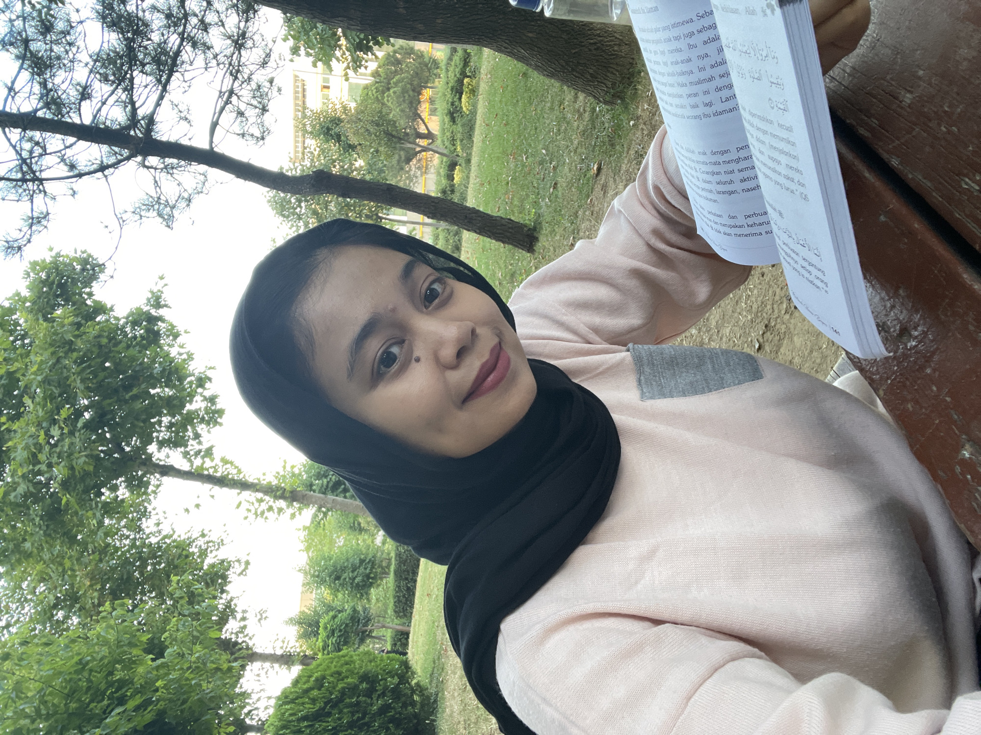 A young woman wearing a black hijab and a light pink sweater is taking a selfie outdoors in a park setting, likely in Başakşehir, Turkey, given the presence of mature trees and manicured green spaces. She is seated at a wooden table, holding an open book with text in Arabic script. The background shows lush greenery, trees, and a building in the distance. The woman is smiling and looking directly at the camera. A partially visible water bottle is on the table to her right. The lighting suggests it is daytime, possibly late afternoon or early evening.