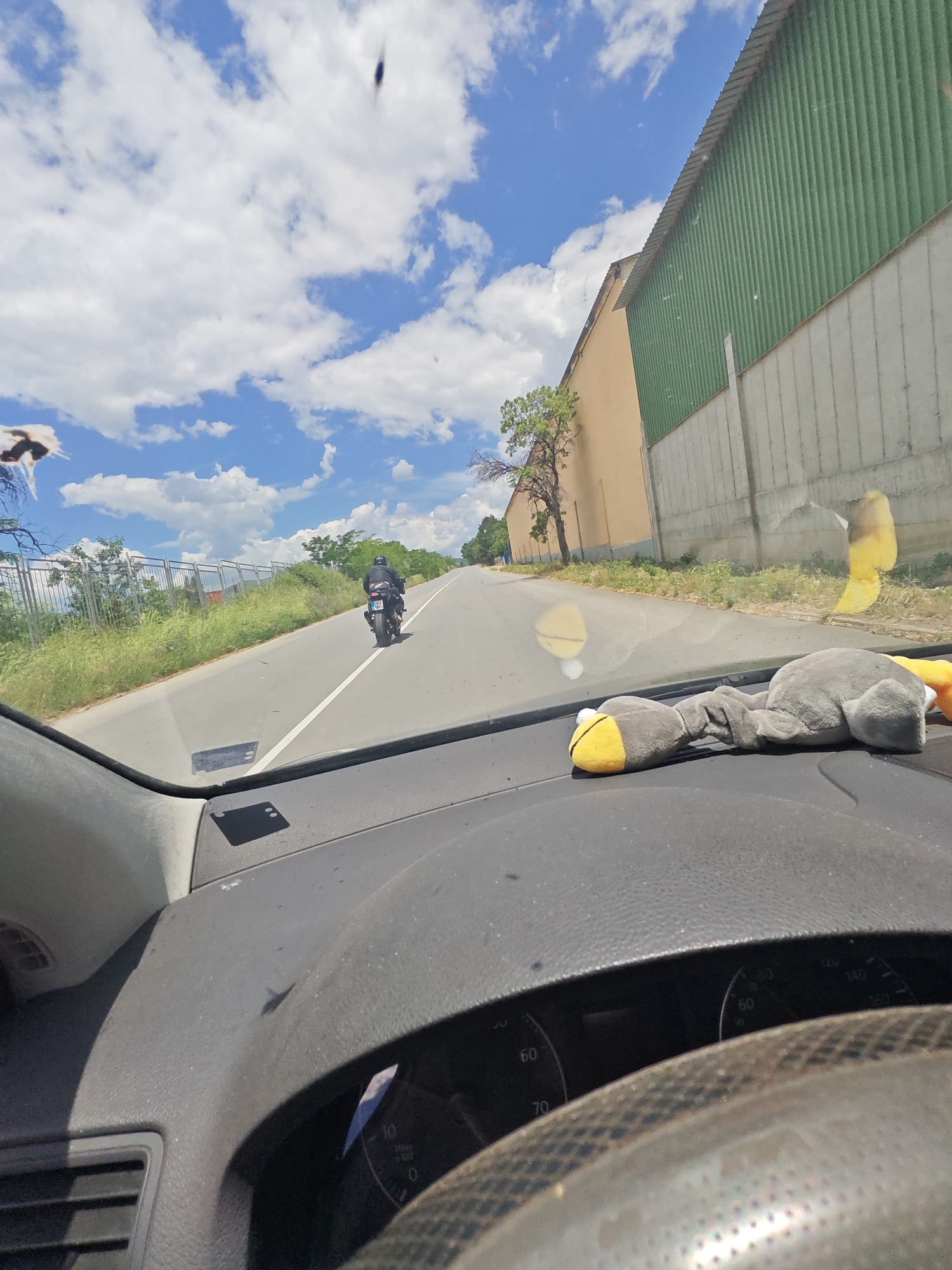 Captured from inside a vehicle, the image shows a road in what appears to be an outskirts area, perhaps near Nova Zagora, Bulgaria. A motorcycle is seen ahead on the road, moving away from the camera. A green and white structure runs parallel to the road on the right. The car's dashboard is visible, with a stuffed animal resting on it. Above, a partly cloudy sky dominates the scene.