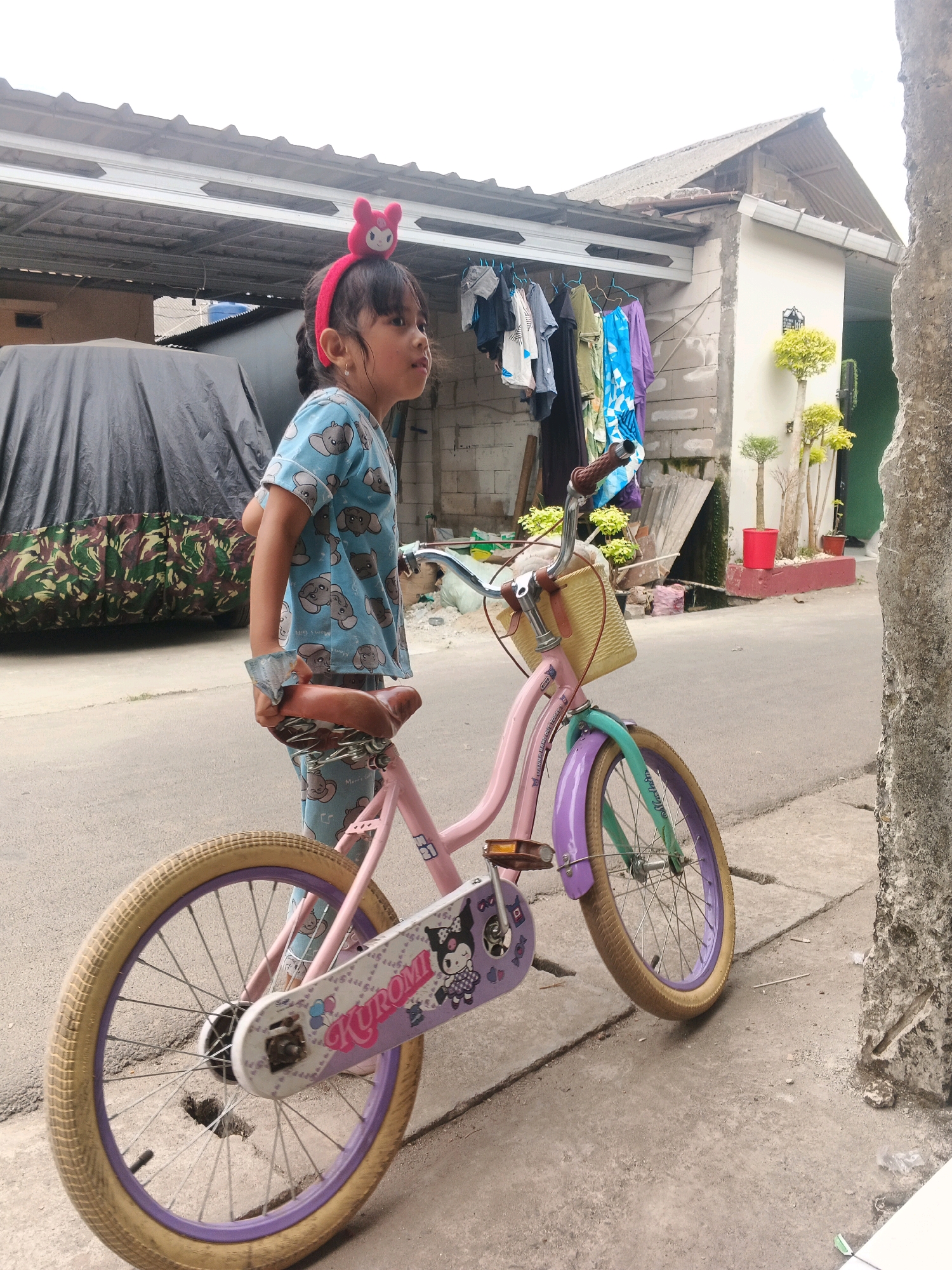 In Kadu, Indonesia, a young girl stands next to a pink bicycle with a yellow basket. She is wearing a blue pajama set with an elephant pattern and a pink headband with a cartoon rabbit character. The bicycle has purple fenders and a purple rim on the rear wheel. A white chain guard features a cartoon character and the word "Kuromi". The girl is holding the bicycle's seat. The background shows a residential area with houses, a covered vehicle, and laundry hanging on a clothesline. The ground is paved with asphalt and concrete.