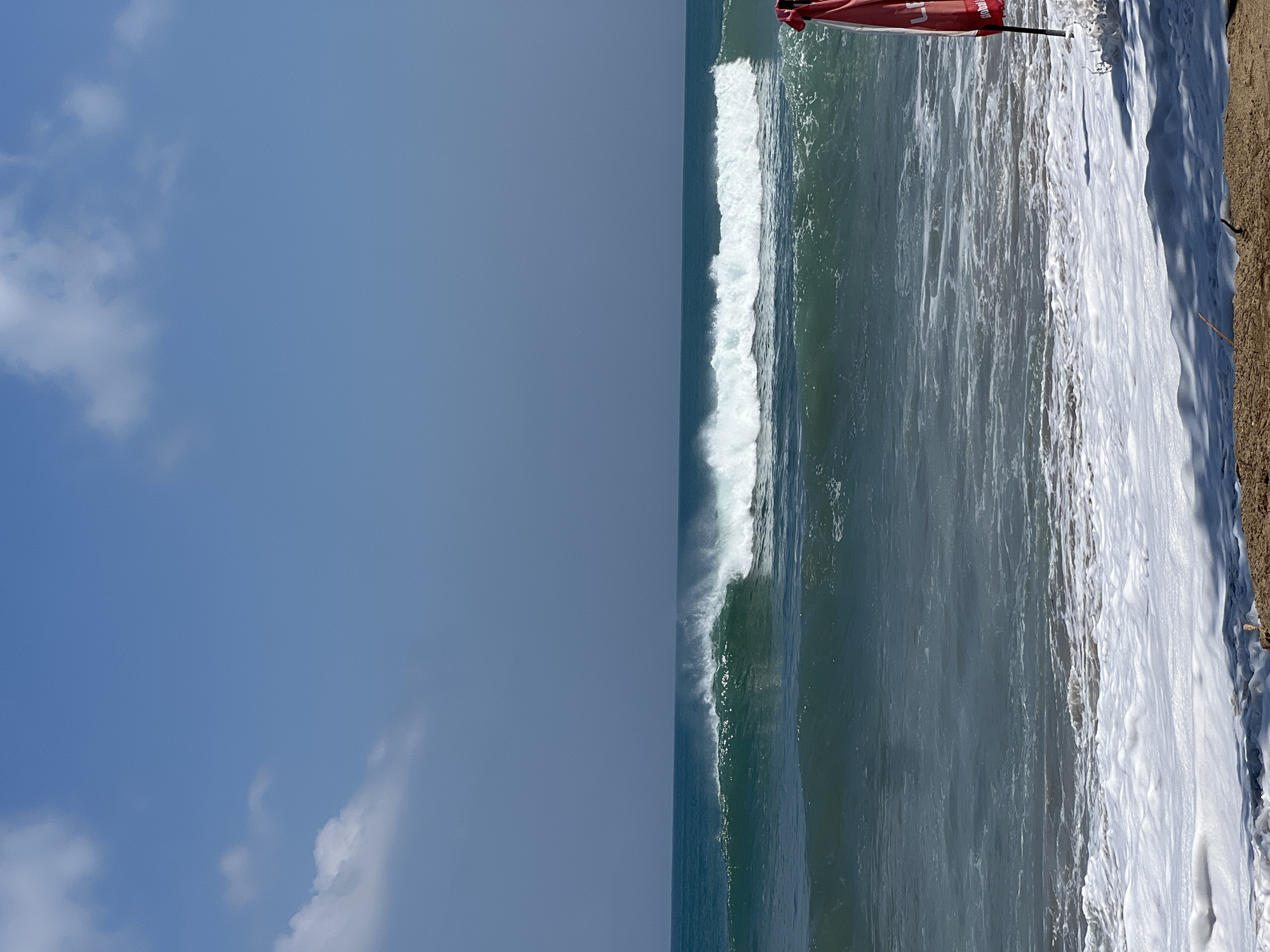 A wide shot captures a section of Kuta beach, Indonesia, on a clear, sunny day. The sky above is a vibrant blue, with a few wispy white clouds. The ocean is a deep turquoise, with a prominent wave breaking in the middle ground, creating a white crest of foam. The foreground is the sandy shore, with white foam from a previous wave receding. A bright red flag, possibly a warning or marker, is visible on the right side of the frame, partially obscured by the foam. There are no people visible in the frame, and the scene is devoid of any human activity. The overall impression is one of a tranquil, natural coastal environment.