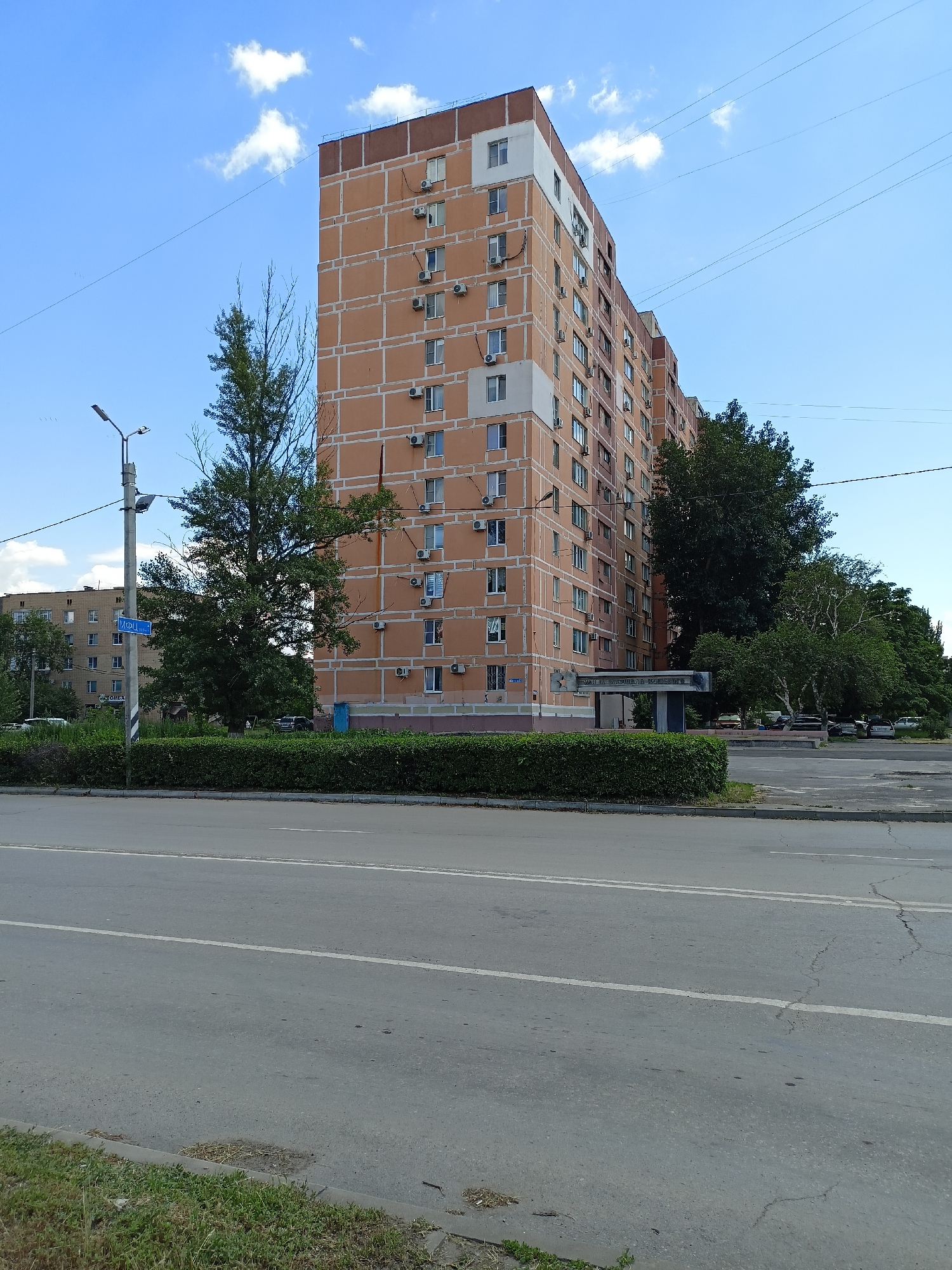 The image depicts a wide asphalt road in front of a multi-story residential building in Volgodonsk, Russia. The building is orange with white trim and has numerous windows and air conditioning units. A light pole with a street sign that reads "ул. Ленина" (Lenin Street) is visible to the left of the building, along with a trimmed green hedge. A large tree stands between the street sign and the building. In the background, more buildings and trees are visible, as well as a parking lot with several cars. The sky is blue with scattered white clouds. The overall scene is that of a typical urban environment on a sunny day.