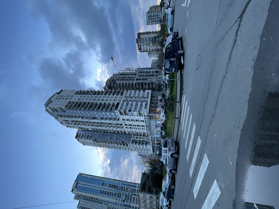 The image depicts an urban street scene in Batumi, Georgia, under an overcast sky, likely during late afternoon or early evening. The foreground features an asphalt road with visible cracks, a white pedestrian crossing (zebra stripes) on the left, and a large puddle reflecting the sky and buildings on the lower left.
In the midground, multiple vehicles are present. On the left, a silver SUV and a dark gray sedan are parked parallel to the curb. Towards the right, a dark blue SUV is positioned near an intersection or roundabout with a small, grassy central median containing sparse vegetation. Other vehicles are visible further down the street in both directions, some parked and others appearing to be in motion around the intersection. A street light pole with a single luminaire stands on the right side of the intersection. A circular manhole cover is visible in the asphalt near the center-right.
The background is dominated by several high-rise residential or mixed-use buildings. The most prominent structures are two tall, modern apartment blocks with light-colored (white/gray) facades and numerous balconies, some with blue-tinted windows. Smaller, less distinct buildings are visible further in the distance. The ground floors of the main buildings appear to house commercial establishments. No distinct individuals are clearly visible. The sky is largely cloudy, transitioning from a lighter blue near the horizon to darker gray clouds overhead, with a hint of brightness in the center-right suggesting a break in the clouds or the setting sun behind them.