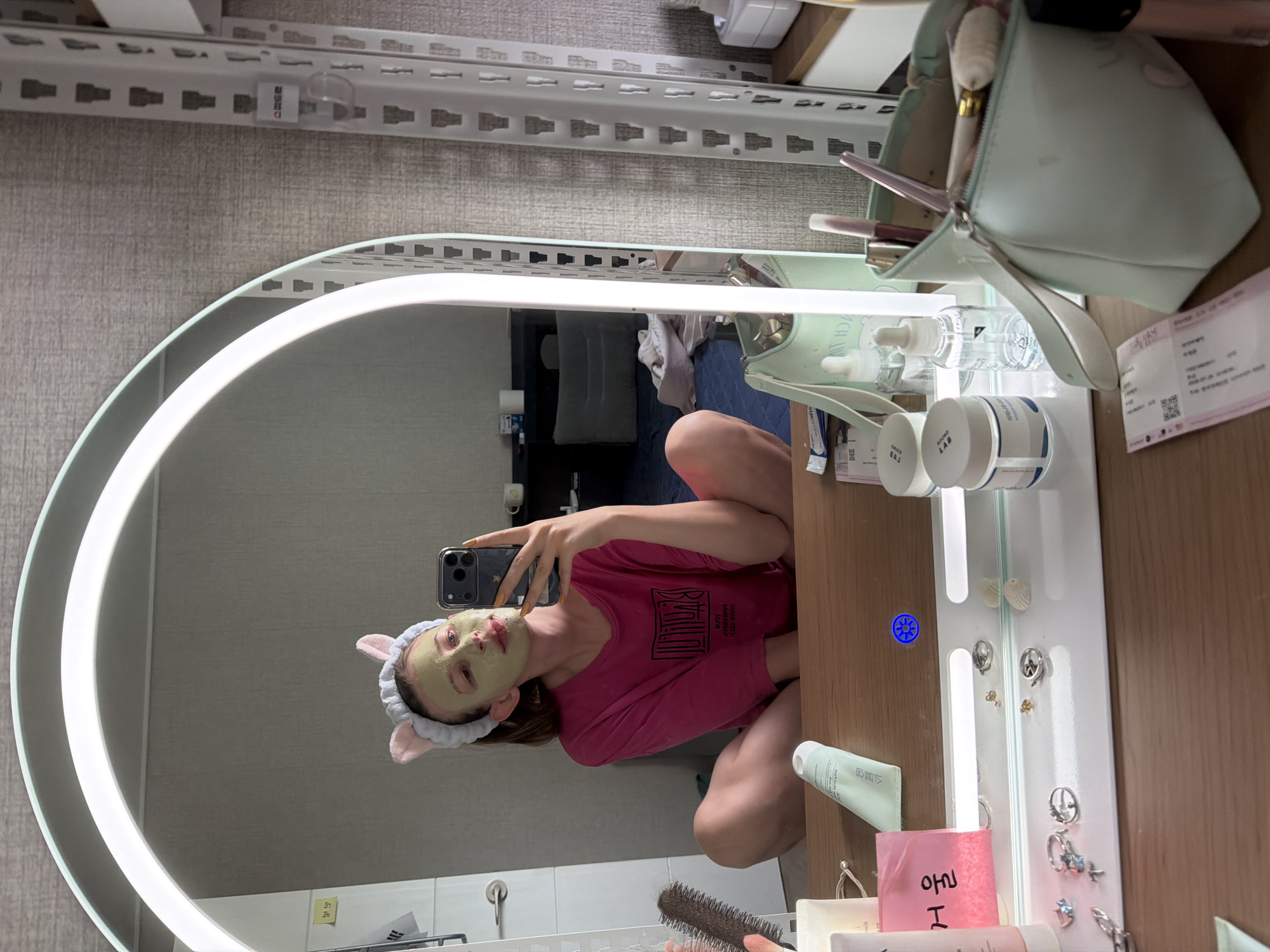 In Dongducheon-si, South Korea, a young woman is taking a selfie in a mirror. She is wearing a pink t-shirt and a cat-ear headband. Her face is covered in a green face mask, and she is holding a smartphone in her right hand. The background shows a bedroom with a bed, a dresser, and a closet. The mirror is large and oval-shaped with a light ring around it. On the dresser, there are various cosmetic items, including a makeup bag, jars, and bottles. The overall scene suggests a moment of self-care and personal grooming.