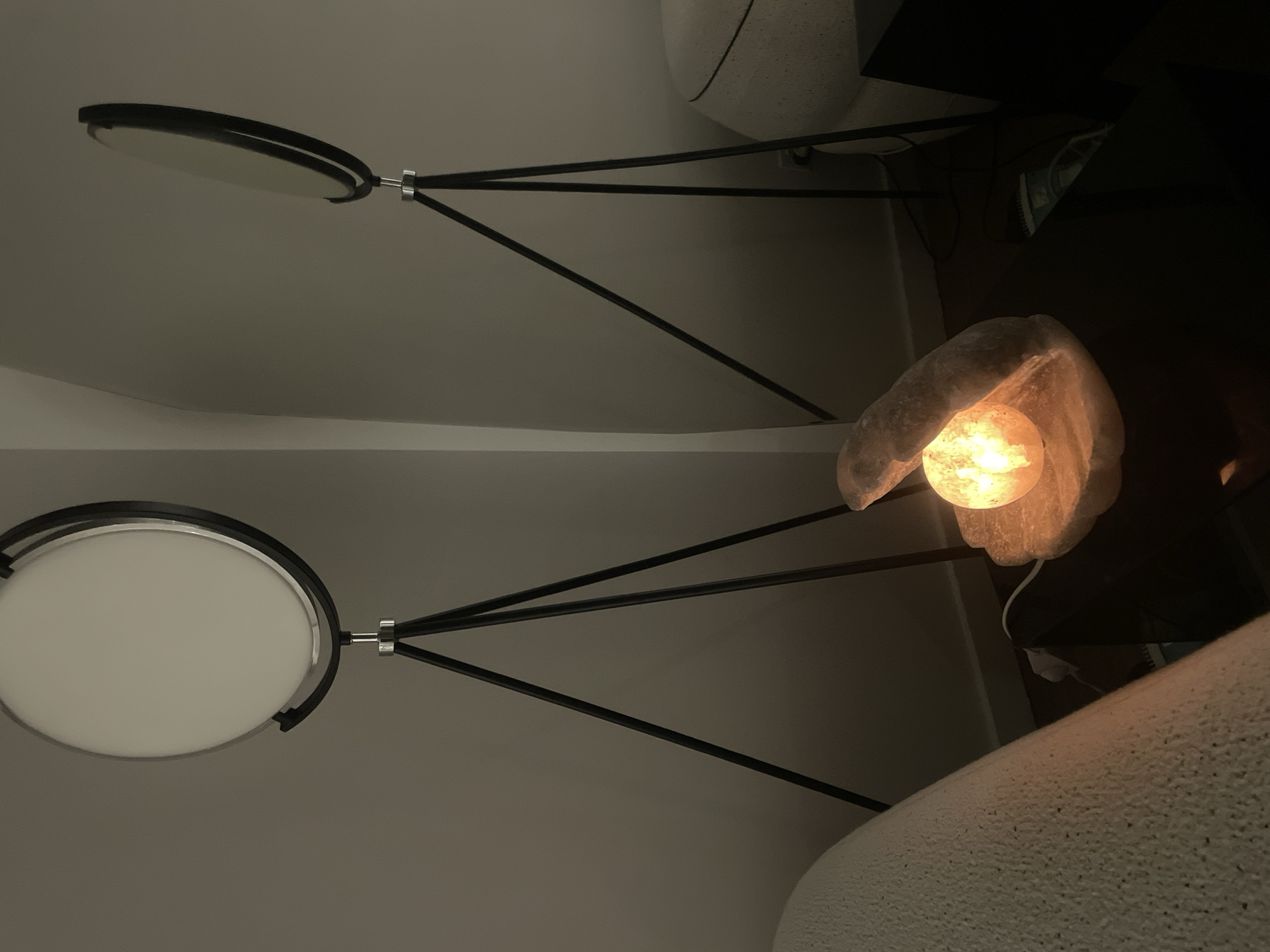The image depicts an indoor scene with ambient lighting, likely a bedroom or living area. Two tripod floor lamps with circular white shades are visible. In the foreground, a salt lamp shaped like a shell with a glowing orange orb sits on a dark surface. A portion of a light-colored textured blanket or sofa is in the lower left corner. The background is a plain wall, with a slight curve in the upper right. No people or animals are present. The location is not specified as being in Brisbane, Australia.