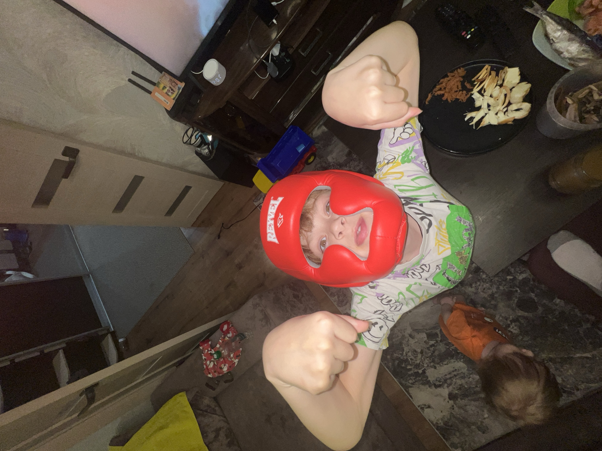 A child wearing a red boxing headgear is positioned in the center of the image, with arms raised in fists. A younger child is lying on the floor near the child with the headgear. The floor is patterned with a marble-like design. On a nearby table, there is a plate of food including what appears to be dried fish and other food items in containers. In the background, there is a TV stand with electronics and toys. The precise location in Noviy, Russia is not discernible from the image.