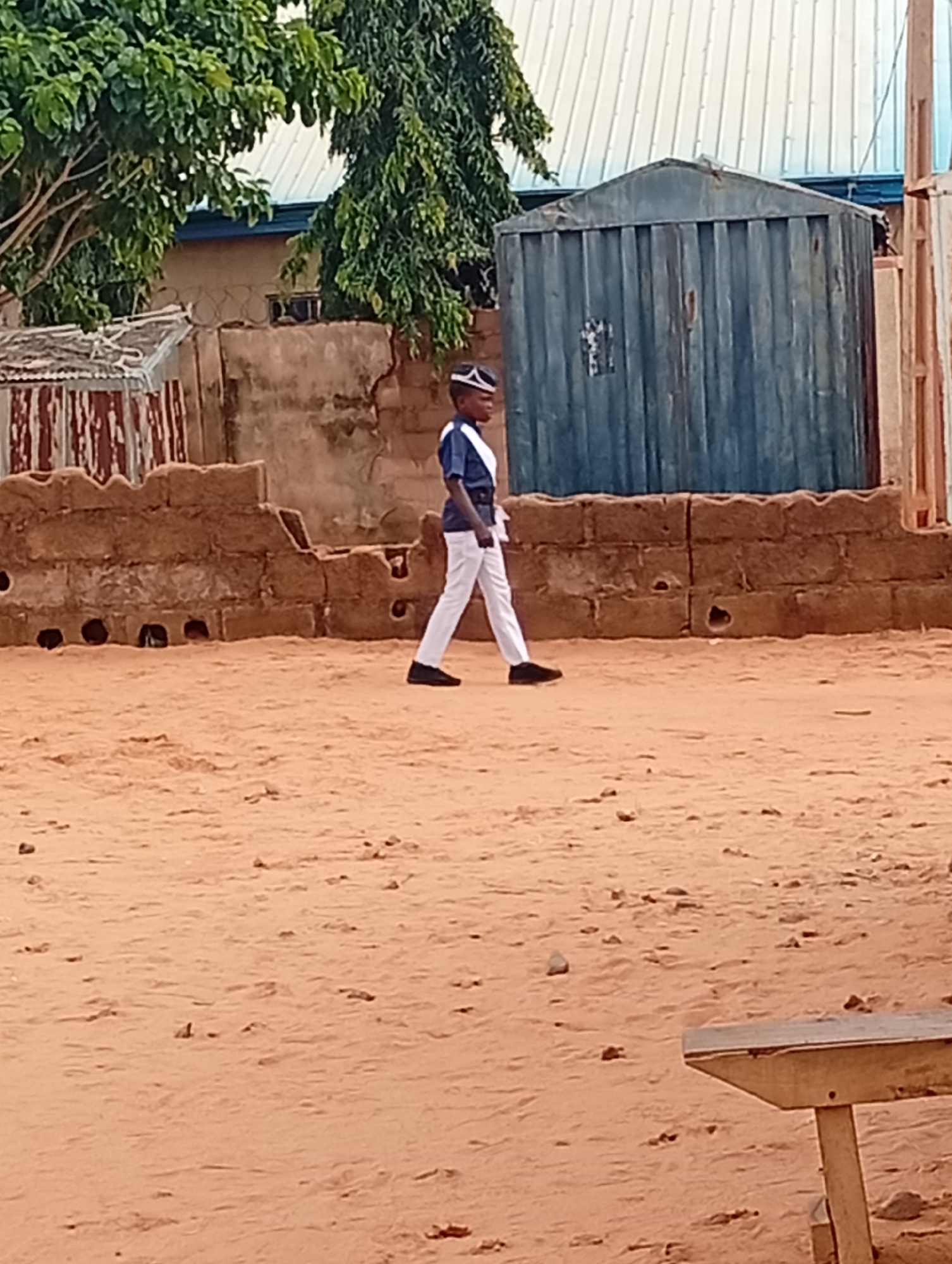 A full shot shows a young person in uniform walking from left to right across a dusty, sandy area. The person is wearing a blue shirt with white trim and a white hat with a black brim, along with white pants and black shoes. Behind them, a low, rough brick wall with openings runs across the mid-ground. Further back, a dark blue metal container with vertical ridges stands to the right of the frame. The background features a building with a white corrugated metal roof, and some trees with green foliage are visible on the left side. A portion of a wooden bench is in the lower right corner of the image.