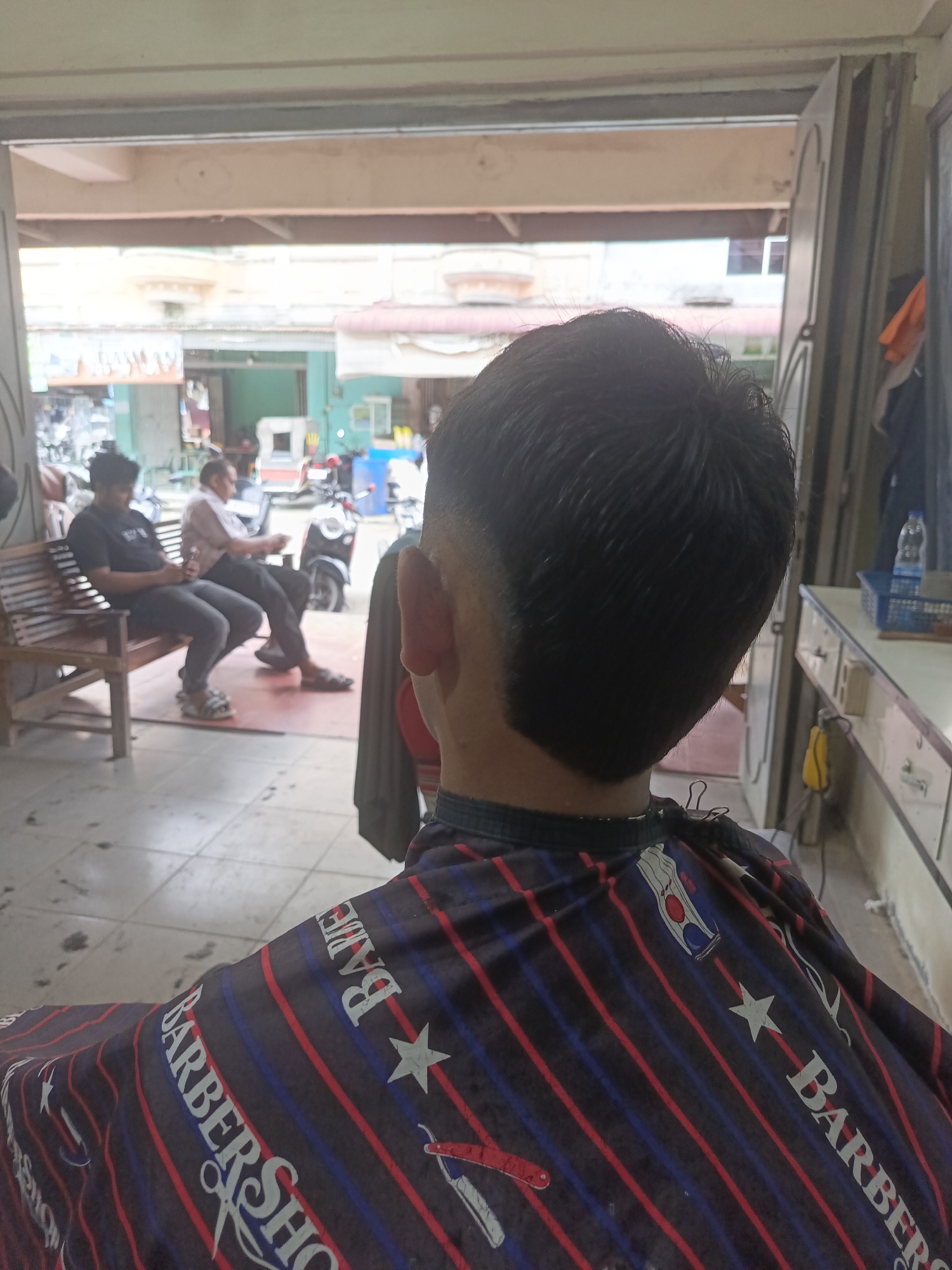 The image depicts the interior of a barbershop in Lhokseumawe, Indonesia. The perspective is from behind a person seated in a barber's chair, wearing a barber's cape. The cape is patterned with diagonal red and blue stripes, interspersed with white stars and the words "BARBER SHO" visible in large white font.
In the background, two men are seated on a wooden bench. The man on the left is wearing a dark t-shirt and jeans, looking down at his phone. The man on the right is wearing a light-colored patterned shirt and dark pants, also looking down. Several motorcycles are visible parked outside the open doorway of the shop, suggesting a street-level location. The floor is tiled, with some dark specks possibly indicating hair clippings. A counter with drawers and a blue basket containing items is visible to the right. The overall lighting suggests it is daytime.