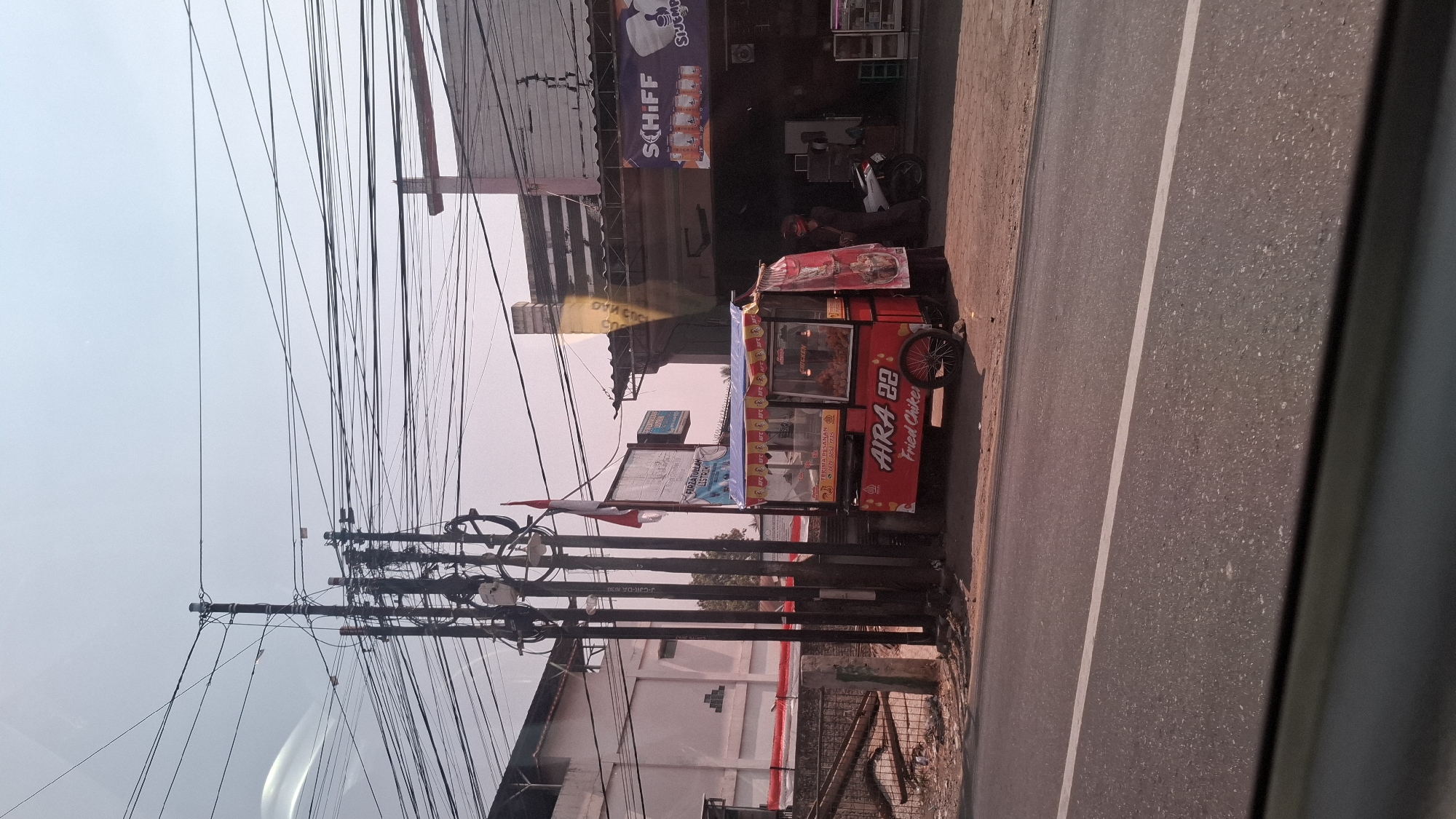 The image captures a street scene in Sirnagalih, Indonesia, dominated by an overhead network of electrical wires and utility poles. A red food cart, branded "AIRA 22 Fried Chicken," is prominently displayed on the sidewalk. The cart has glass displays showcasing fried chicken and a banner that reads "TERIMA PESANAN" (Take Orders). A woman in dark clothing, wearing a helmet, stands near the cart, suggesting she might be the vendor or a customer. A white motorcycle is parked behind the food cart. To the right of the cart is a building with a signage that says "SCHIFF." In the background, there are other structures and a tall utility pole with an Indonesian flag attached. The ground is a paved road with a white line marking a lane. The sky is a pale blue, indicating daylight.
