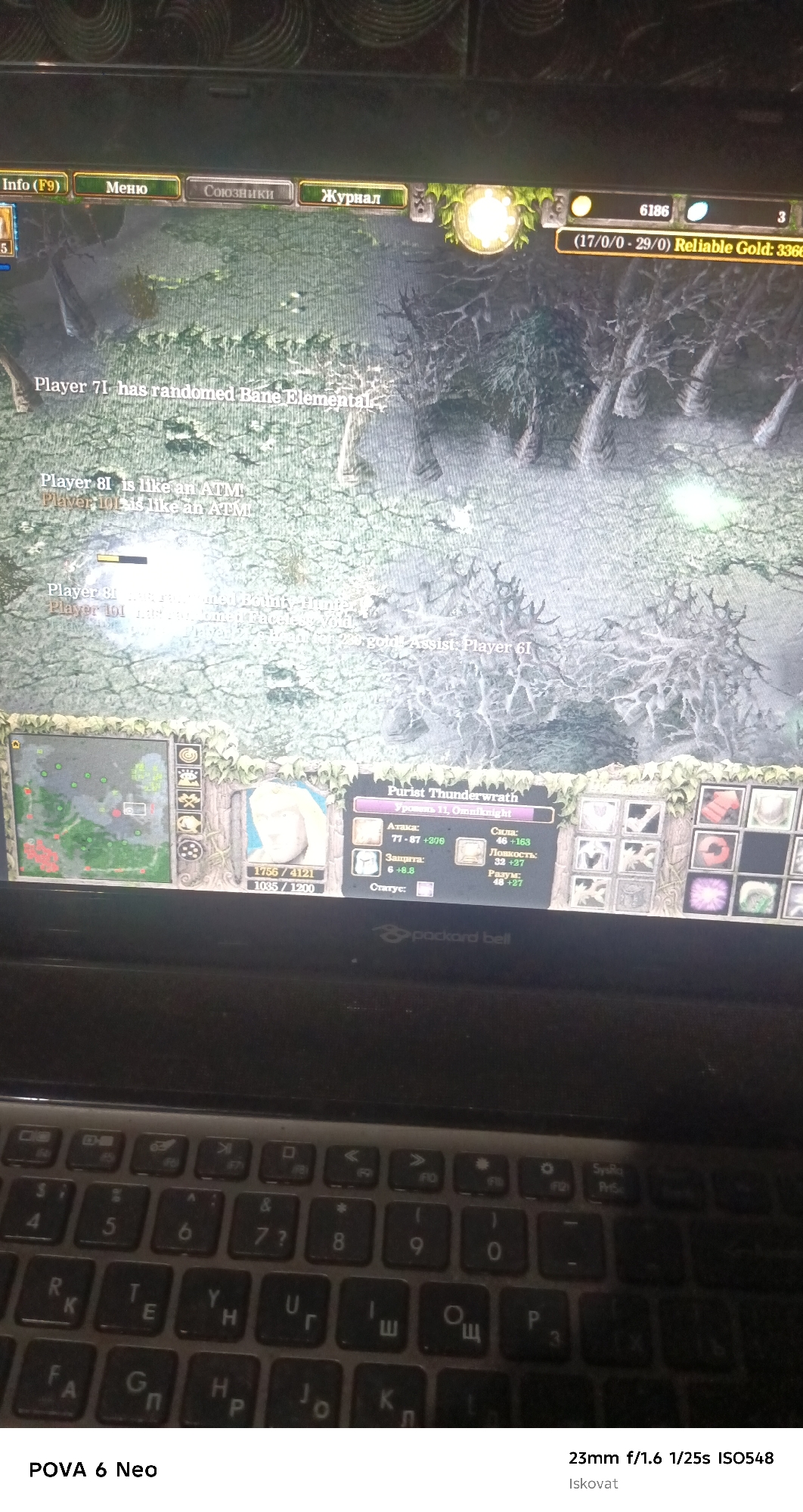 The image shows a laptop screen displaying a video game, likely Warcraft III given the interface elements and unit portraits. The game appears to be in the midst of a match, with player names and statistics visible. The laptop is placed on a surface, and a keyboard is partially visible in the foreground. The background of the laptop screen depicts a dark, forested environment, characteristic of a fantasy setting. The surrounding environment, including the texture on the laptop lid, suggests an indoor setting. The text at the bottom indicates the camera's settings and the device used, "POVA 6 Neo" and "23mm f/1.6 1/25s ISO548". There is no visible indication of the city of Pastki-Peshkurgan, Uzbekistan, within the image itself.