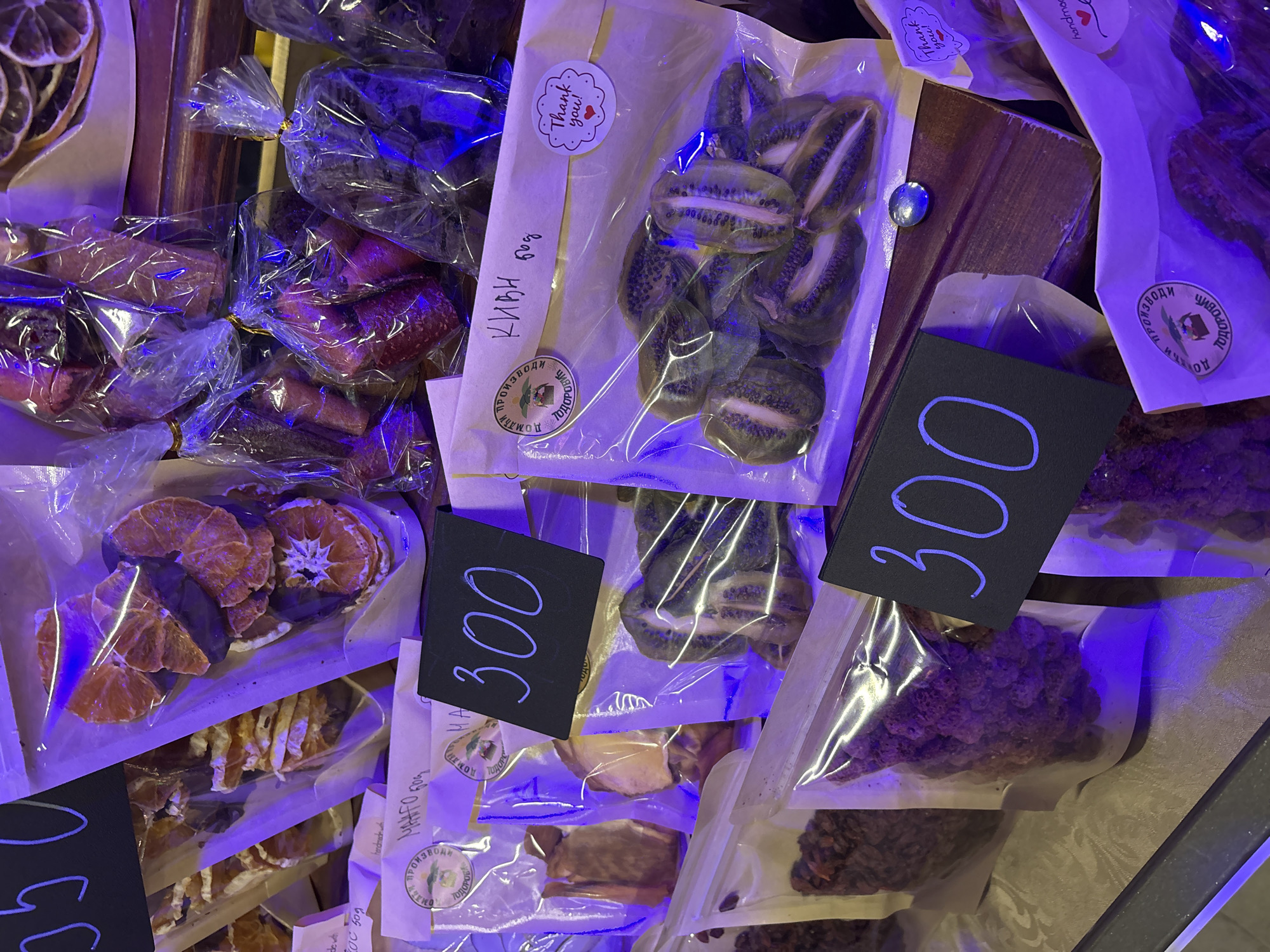 The image shows a display of packaged dried fruits in Kragujevac, Serbia. Clear plastic bags contain various fruits, including sliced oranges, kiwi, and other unidentified dried fruit. Black price tags with "300" are placed near some packages. Each package has a white label with the brand name and other information. The scene is lit with a strong blue light.