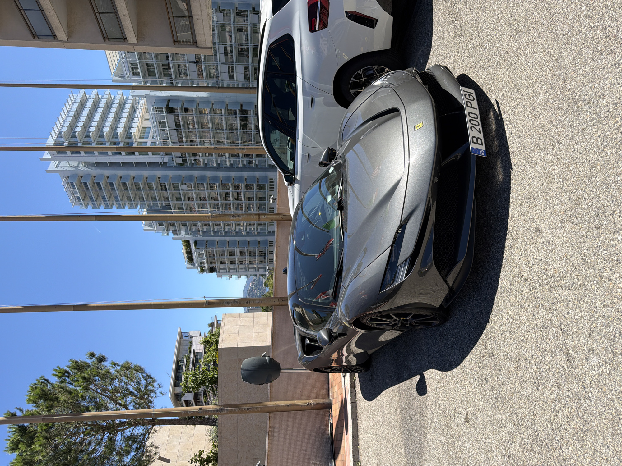 A gray Ferrari SF90 Stradale is parked on a paved surface in front of modern buildings in Monaco. The car is facing slightly to the left, with its front bumper and hood prominently displayed. The license plate, "B 200 PGI", is visible on the front. To the right of the Ferrari, a white SUV is partially visible. In the background, tall, white apartment buildings with balconies and glass facades dominate the skyline. A large green tree is situated to the left of the Ferrari, with a beige building behind it. The sky is clear and blue, suggesting a sunny day.