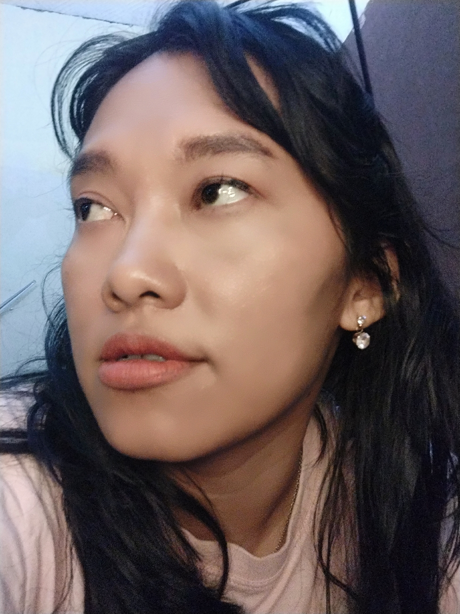 This is a close-up, selfie-style photograph of a young woman with long, dark hair and fair skin. She is looking off to the left, with her head tilted slightly. She is wearing a light pink shirt and a delicate earring with a clear stone. The background is blurred, with a light blue wall on the left and a darker, textured surface on the right. The lighting appears to be artificial. The image does not provide any visual information that would allow for identification of the location within Tasikmalaya, Indonesia, or any specific environmental context.