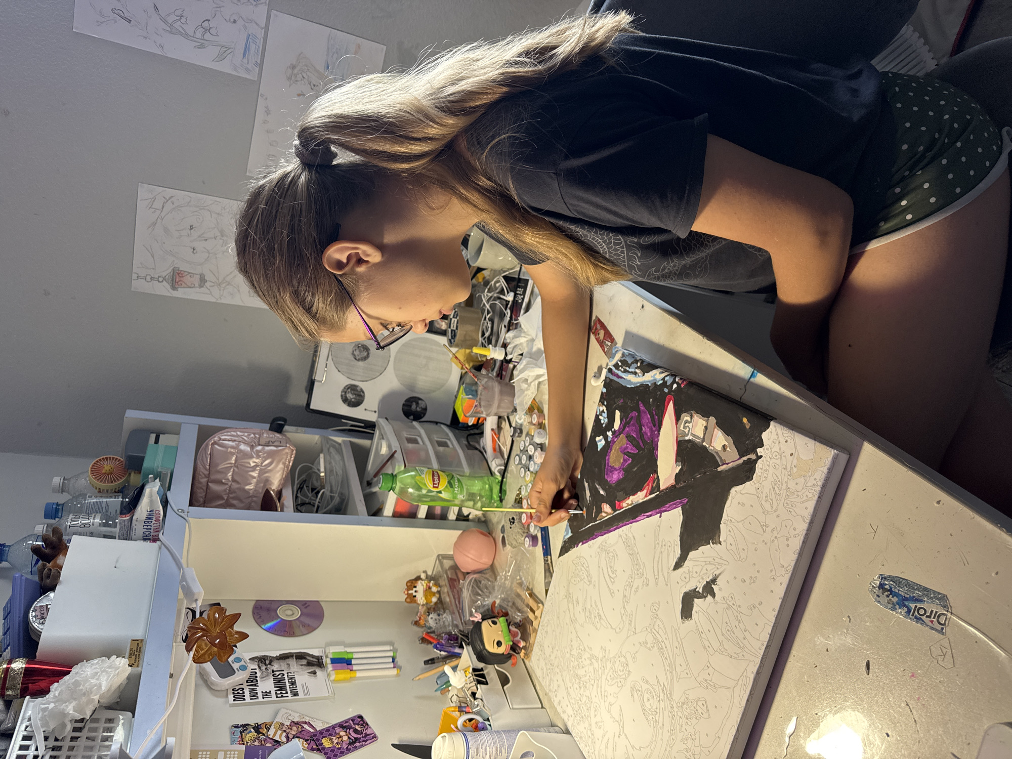 A young girl with blonde hair pulled back in a ponytail and wearing glasses is painting at a desk. She is wearing a dark t-shirt and green polka dot shorts. The desk is covered with art supplies, including paint pots, brushes, and a paint-by-numbers canvas. The canvas appears to be a portrait with black and purple accents. The background shows a shelf filled with various items, including water bottles, a pink makeup bag, and drawings taped to the wall. The scene appears to be an indoor art studio or a bedroom.