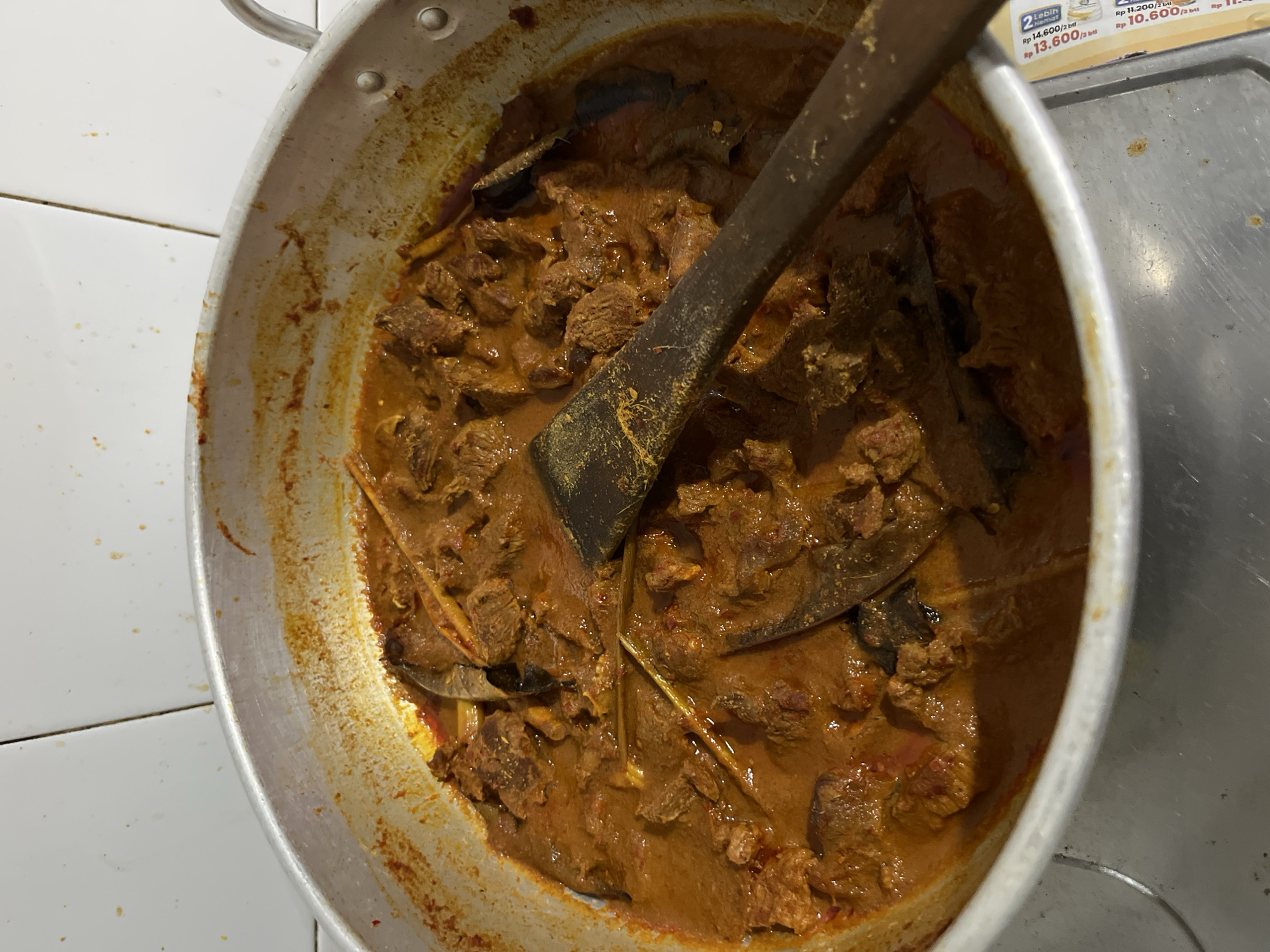 The image shows a metal pot filled with a dark orange/brown stew-like dish, likely a curry or similar savory dish featuring chunks of meat and various spices. A dark-colored, possibly wooden, spatula rests within the pot, partially submerged in the food. The pot sits on a stovetop with what appears to be pricing/promotional information from a store. The location is likely Keudematangkuli, Indonesia, although this cannot be confirmed from the image itself. The focus is on the food preparation and a meal, but without further details, the exact ingredients or preparation method cannot be determined. The environment appears to be a kitchen setting.