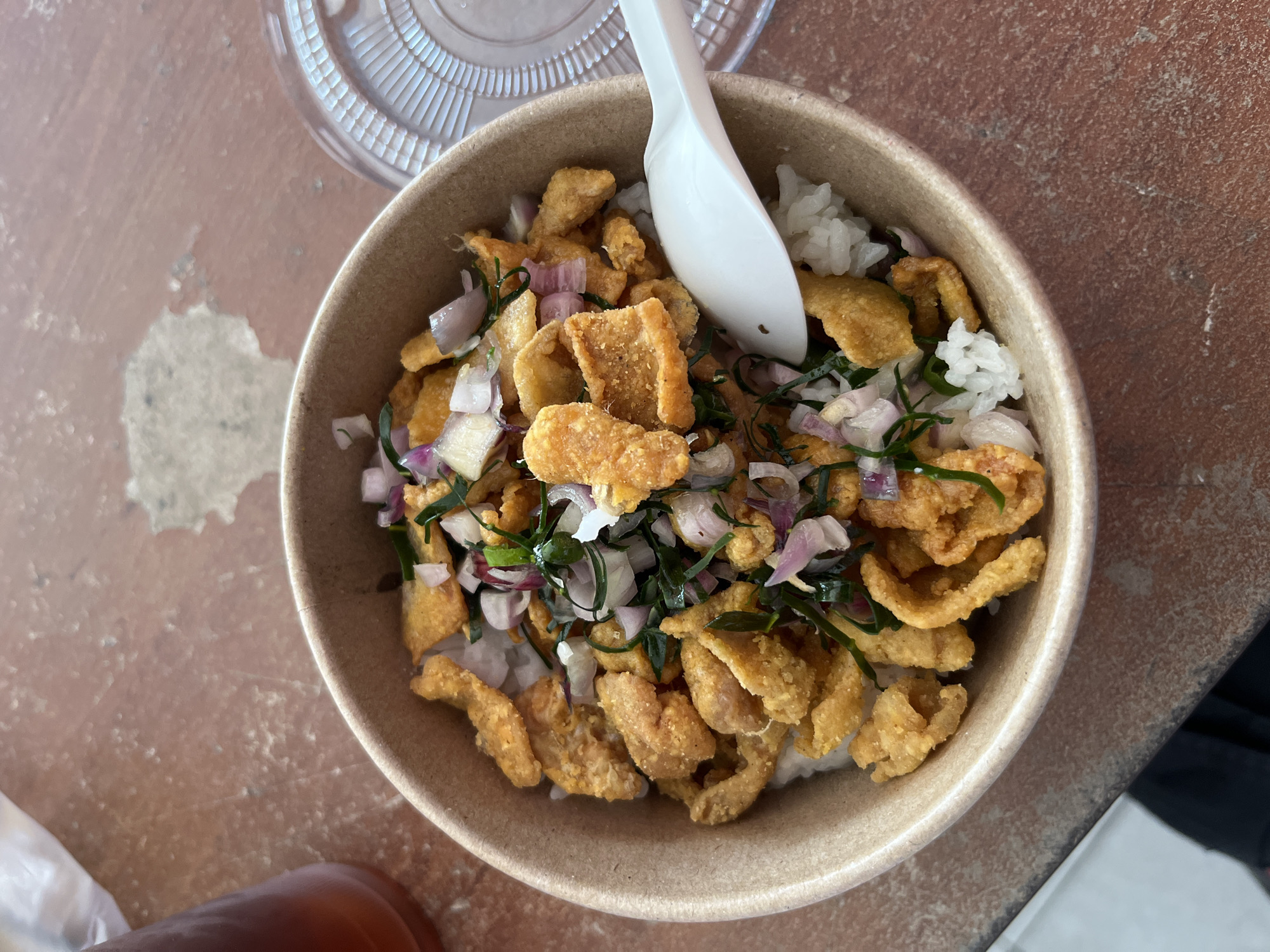 A bowl of rice topped with fried chicken skin pieces, sliced red onions, and green herbs sits on a reddish-brown textured surface. A white plastic spoon rests on the edge of the bowl. To the left, a portion of a dark beverage in a clear plastic cup is visible, and to the right, a clear plastic lid for the bowl is partially seen. The scene is captured from a top-down perspective.