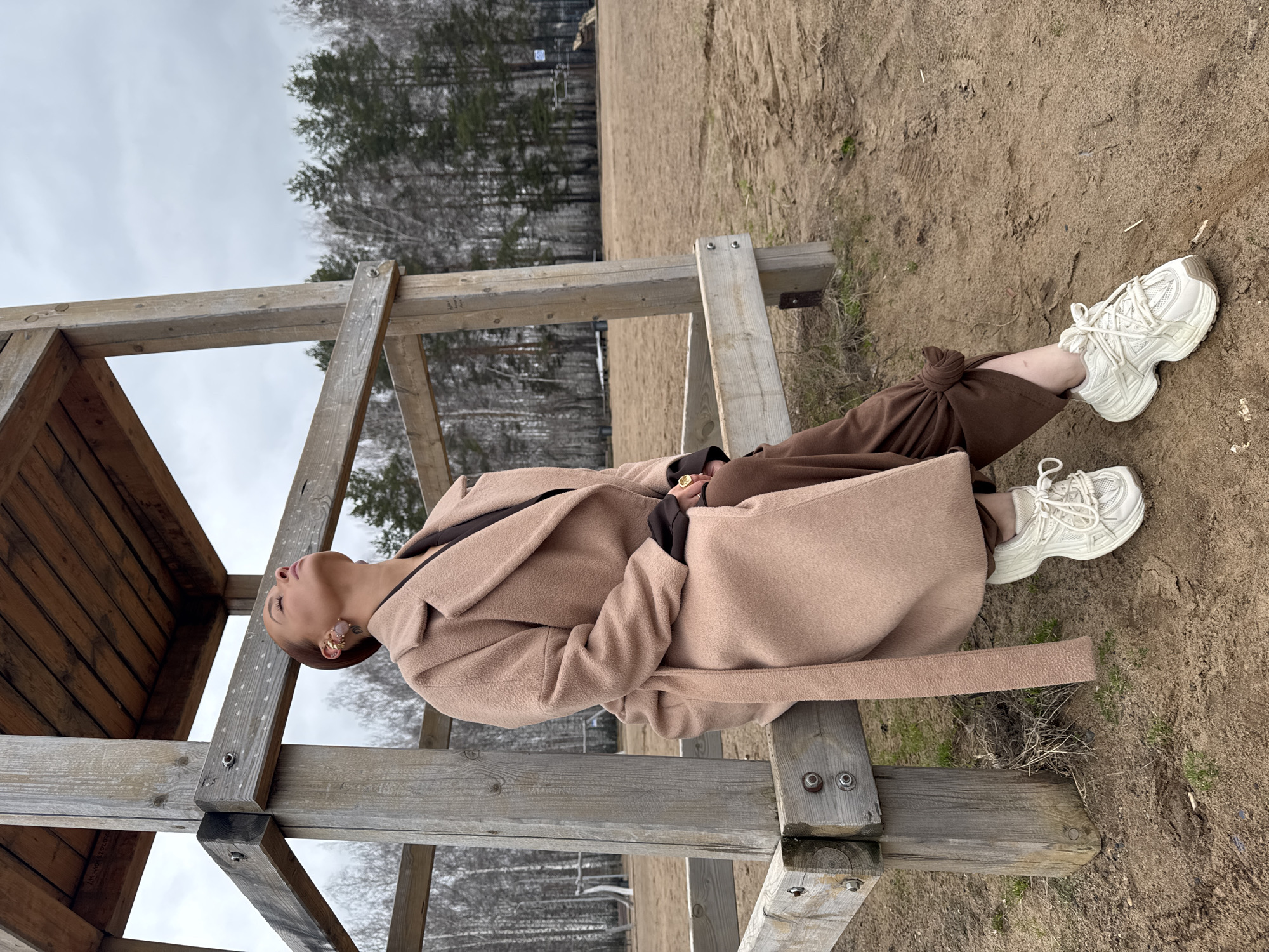 The image shows a woman reclining on a wooden structure in what appears to be an outdoor environment. She is wearing a beige coat and brown pants, complemented by white sneakers. The backdrop features trees and a sandy terrain, suggesting a park or natural area. Based on additional information, the location is likely in the city of Chaikovsky, Russia.