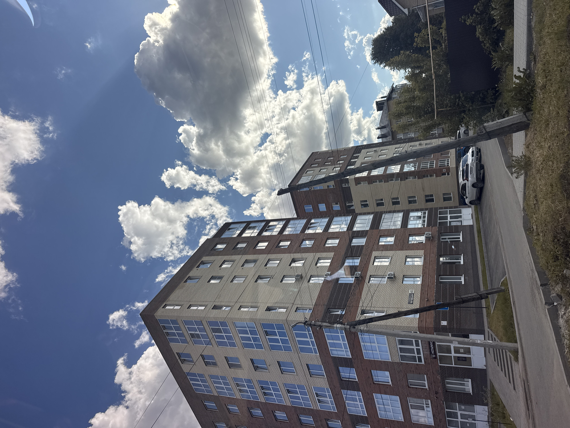 The image captures a street scene in Mozhga, Russia, dominated by two multi-story apartment buildings. The buildings are constructed with a combination of light-colored brick and darker, reddish-brown brick. The facades are punctuated by numerous windows, many reflecting the clear blue sky with scattered cumulus clouds. Overhead power lines stretch across the sky and intersect with the buildings. A street runs along the side of the buildings, with parked white vehicles and foliage lining the edge. A utility pole with attached street signs is visible near the sidewalk, and signage is visible on the ground floor of one of the buildings. The overall impression is of a sunny, urban residential area.