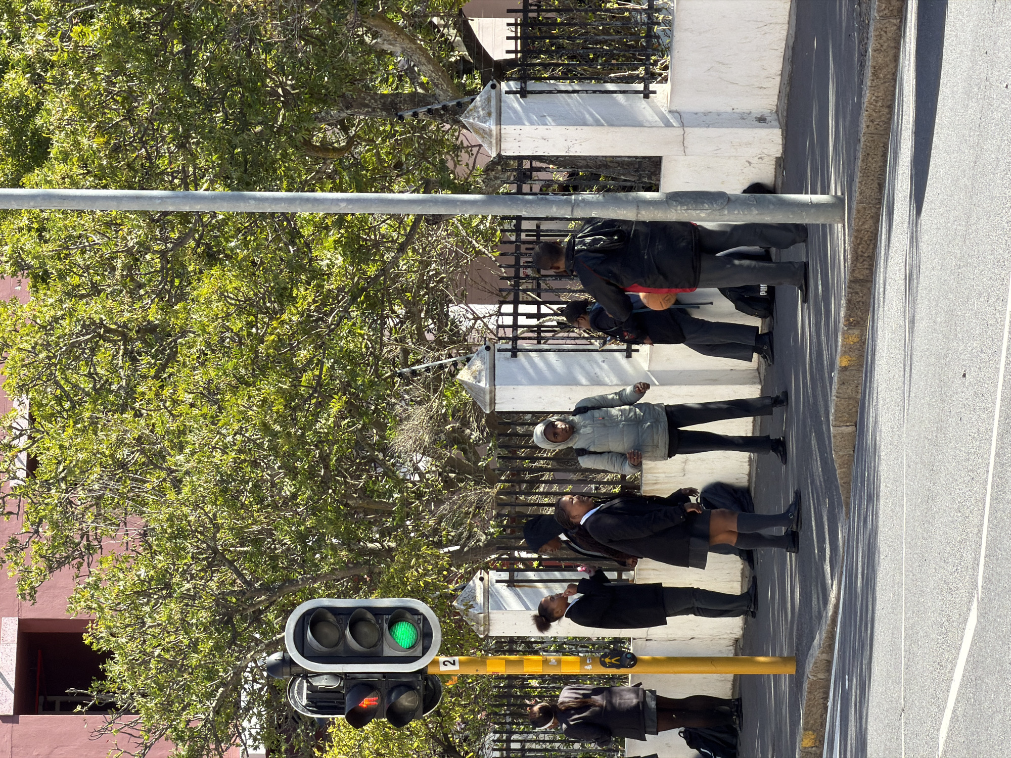 The image depicts a bright, sunny daytime scene on a street in Cape Town, South Africa, featuring several people, likely school students, waiting or walking along a sidewalk. The photograph is oriented vertically, but depicts a horizontal street scene.
In the foreground and midground, a group of approximately seven to eight individuals is visible. Most appear to be young people dressed in dark school uniforms, consisting of dark blazers or jackets, white shirts, and dark skirts or trousers. One person is wearing a light grey hooded jacket. They are standing on a paved sidewalk next to a curb and a road. Distinct, long shadows are cast across the sidewalk and road, indicating clear weather and a sun high in the sky, possibly morning or afternoon.
A traffic light assembly is prominent in the left foreground. The main vehicle traffic light shows a green signal, while a smaller pedestrian signal below it displays a red light, indicating that vehicles have the right of way and pedestrians should wait. A yellow pole with the number "2" written on it is also visible beneath the traffic light.
To the left of the people is a lush green tree with dense foliage, its branches extending over parts of the scene. Behind the people, there's a dark metal fence with white concrete pillars, suggesting a property boundary. A reddish-brown building is partially visible through the tree branches in the background. The street surface shows asphalt with visible white lane markings and a yellow curb.