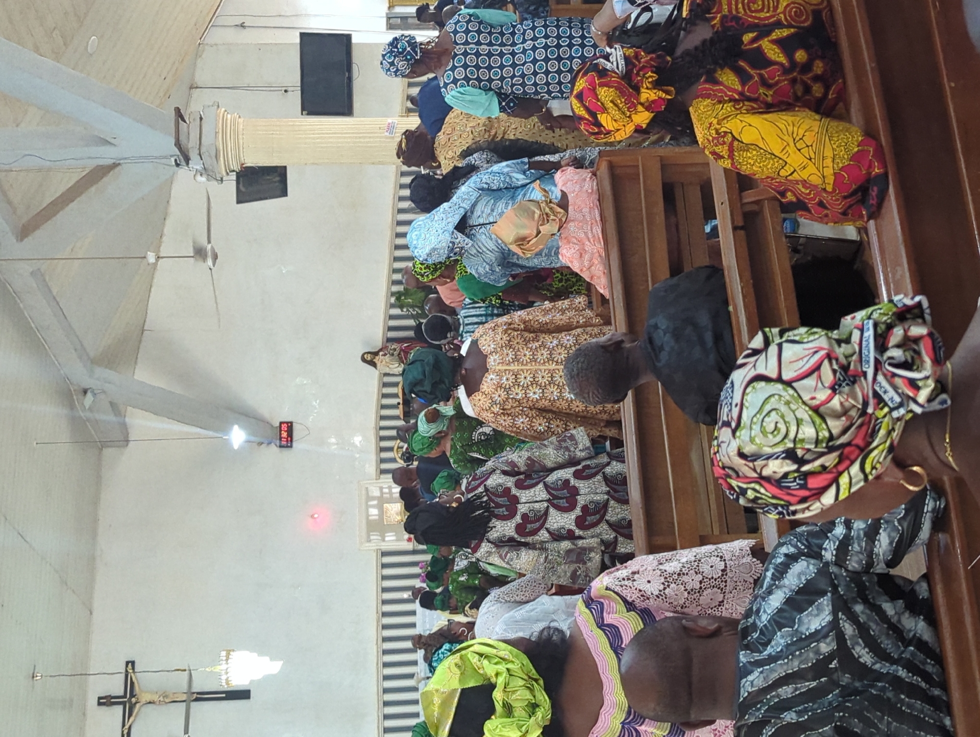 In Narayi, Nigeria, a congregation gathers inside a church. The church's interior features a white, textured ceiling with visible beams and lighting fixtures. A prominent crucifix hangs on the left wall, accompanied by a decorative chandelier. A digital clock displaying "11:32:05" is mounted on the wall near a red light. Two flat-screen televisions are visible on the right side of the church.
The congregation consists of numerous individuals, predominantly women, dressed in vibrantly colored traditional attire and headscarves. They are seated on wooden pews, facing forward, suggesting they are participating in a service or event. Some individuals are standing in the background. The atmosphere appears to be serene and respectful.