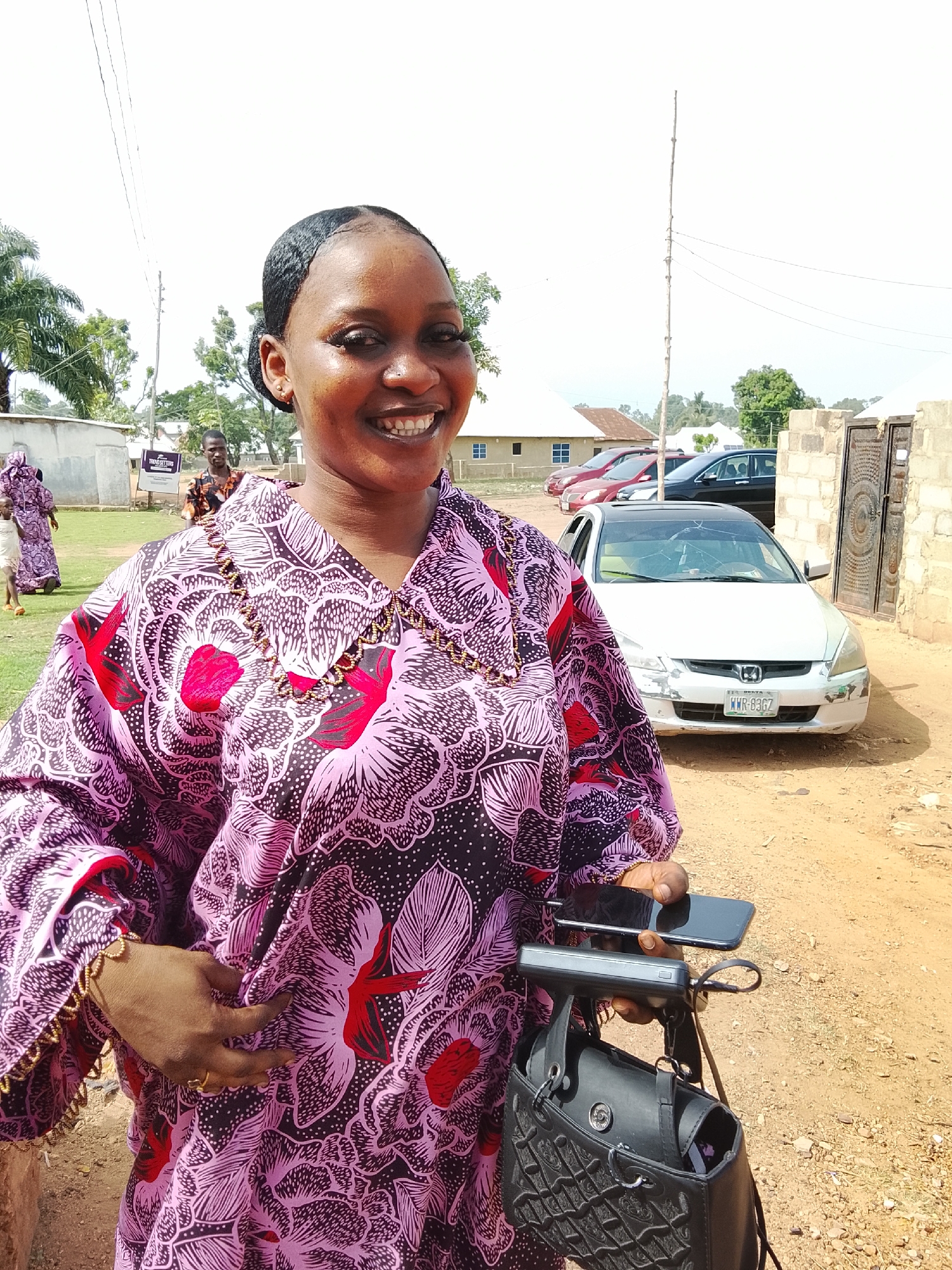 A medium shot captures a woman, likely in her late 20s or early 30s, smiling towards the camera. She is dressed in a patterned pink and black robe-like garment with red floral accents. She holds a smartphone and a black purse in her left hand. The background reveals a rural or suburban scene in Akwanga, Nigeria, evidenced by the dirt road, a partially damaged white car with license plate "WWR-8367", and simple buildings with various cars parked nearby. Other people can be seen in the background, suggesting a gathering or event is taking place. The presence of a stone wall and gate indicates a property boundary. An electricity pole is also visible.