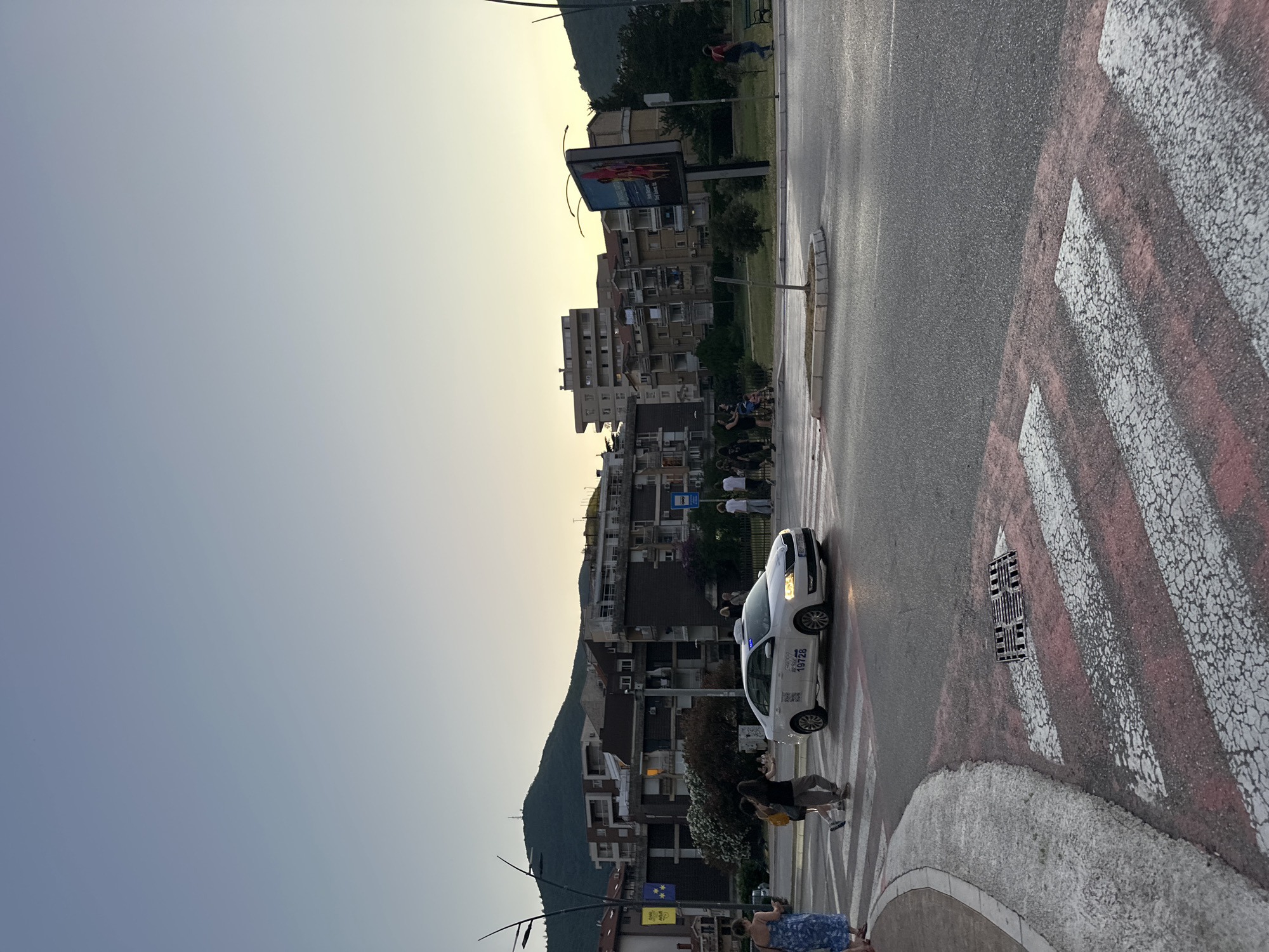 In Budva, Montenegro, a white Skoda Octavia taxi with license plate BD TT 068 is seen crossing a pedestrian crossing on an asphalt road. The taxi has "19728" prominently displayed on its side. Several pedestrians are present, some walking across the street, while others are on the sidewalk. In the background, apartment buildings and a billboard are visible. A tree and a traffic light are also present in the scene. The sky is a clear, pale blue, indicating it is likely late afternoon or early evening. The pedestrian crossing has red and white stripes, with some wear and tear visible on the paint.