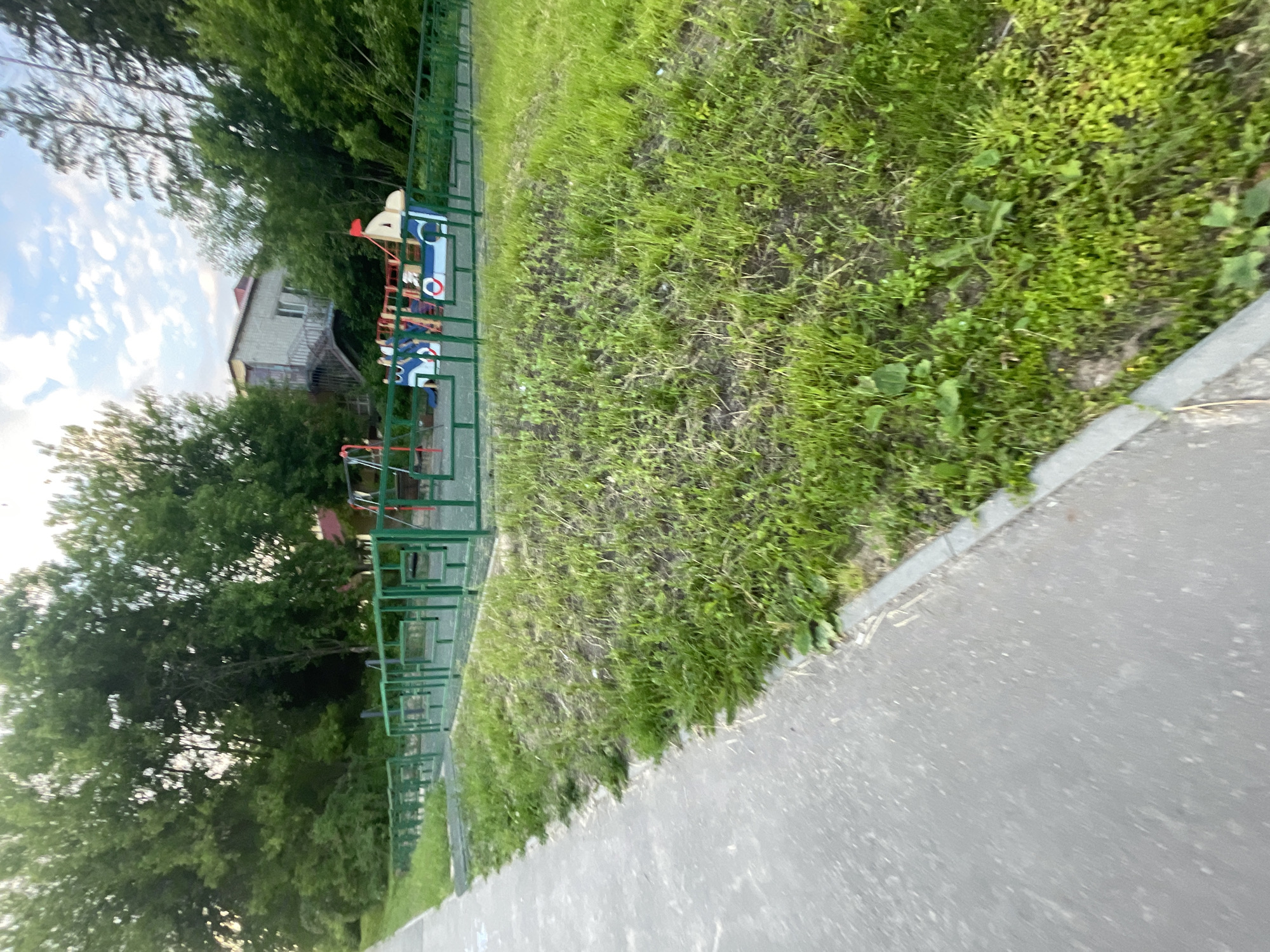 The image shows an outdoor scene, likely in the city of Krasnye Baki, Russia, featuring a playground area adjacent to a paved walkway. The playground is enclosed by a green metal fence and contains several pieces of equipment, including what appears to be a large, boat-shaped play structure with a red flag, and a swing set with red supports. The ground within the playground appears to be a rubberized surface. Surrounding the playground is a grassy area with some patches of dirt. A paved walkway runs along the left side of the image, bordered by a grassy strip. In the background, a two-story building with a light-colored facade and stairs leading to an upper level is visible, partially obscured by dense green trees. The sky above is partly cloudy with a blue and white hue. There are no people visible in the image.