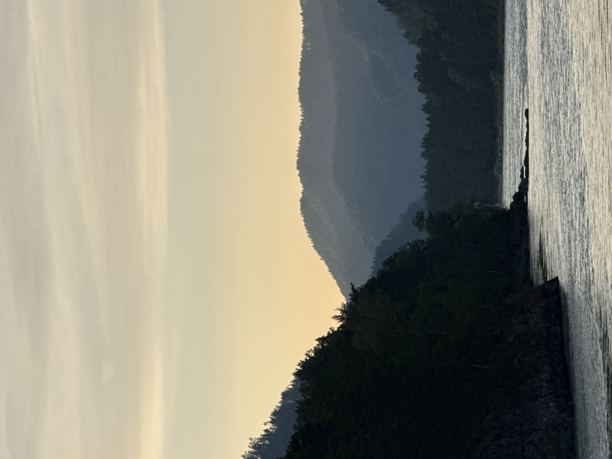 A wide, tranquil river flows through a valley framed by forested mountains. The sky is a soft gradient of pale yellow and pale blue, with a few wispy clouds. On the left, a steep, dark-wooded mountain slopes down to the river, its rocky edge prominent in the foreground. On the right, successive layers of hazy, dark blue mountains recede into the distance, their peaks silhouetted against the sky. The river's surface reflects the light, creating a shimmering, textured appearance. Rocks are visible in the water near the left bank. The scene captures a serene natural landscape at dusk or dawn, likely near Barangol, Russia, given the characteristic mountain and river scenery of the Altai region.