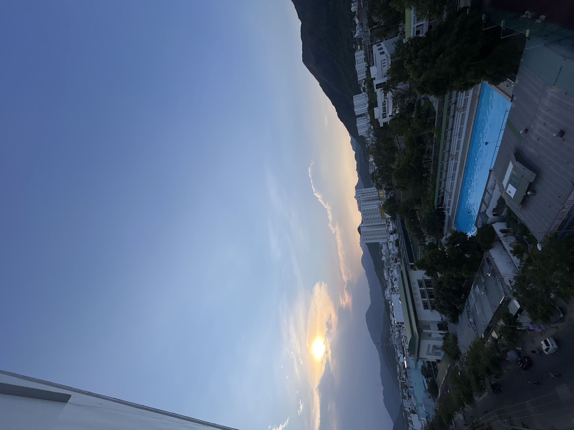 Captured from an elevated perspective, the image presents a landscape of Bac Nha Trang Ward, Vietnam. The sun, partially obscured by clouds, emits a warm glow in the sky. Adjacent to a mountain range are several buildings of varying heights and architectural styles. A long, rectangular swimming pool with blue water can be seen near the buildings. Urban streets, lined with trees and interspersed with cars, are visible in the lower portion of the image. The general ambiance suggests a late afternoon or early evening setting.