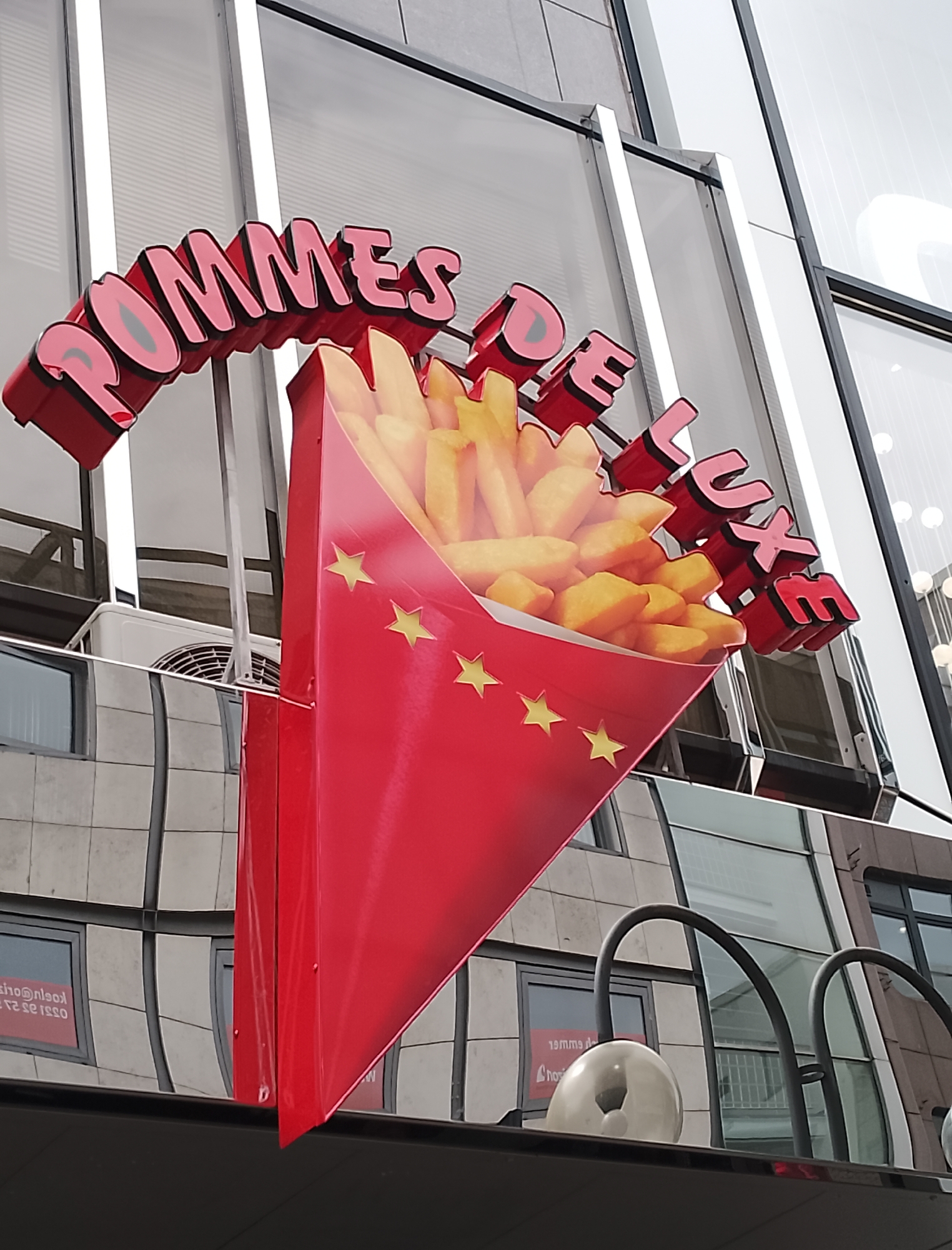 This media file shows an outdoor sign for a french fry establishment. The sign is prominently displayed on a building, featuring a red cone filled with french fries, and the words "POMMES DE LUXE" in large, red, 3D letters. The letters have pink interiors and a black outline. The cone is decorated with yellow stars.
In the background, the building has a modern architectural design with large windows that reflect the sky and surrounding environment, creating a distorted, mirrored effect. An air conditioning unit is visible on the side of the building. The overall lighting suggests it is daytime, with overcast or diffused light.
Reflected in the windows are signs with text that appears to be in a foreign language, possibly French, with some text readable as "19mma de" and "CHOS". The scene is static and focused on the signage, suggesting a commercial setting. There are no people visible in the frame. The image captures a close-up of the sign against the backdrop of the building's facade.