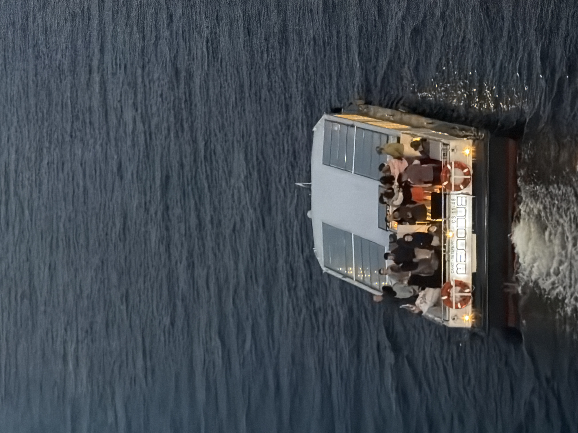 A tour boat, identified by the name "МЕЛОДИЯ" and the registration number "СЗ 11-48" written on its hull, is navigating through the dark waters of a river or canal in Saint Petersburg, Russia. The boat, with a white superstructure featuring glass panels, is carrying a group of approximately 15 passengers who are standing on the open deck, observing the surroundings or each other. The scene appears to be captured during dusk or twilight, as indicated by the low light conditions and the lights illuminating the side of the boat and the water's surface. The boat is creating a wake as it moves through the water.
