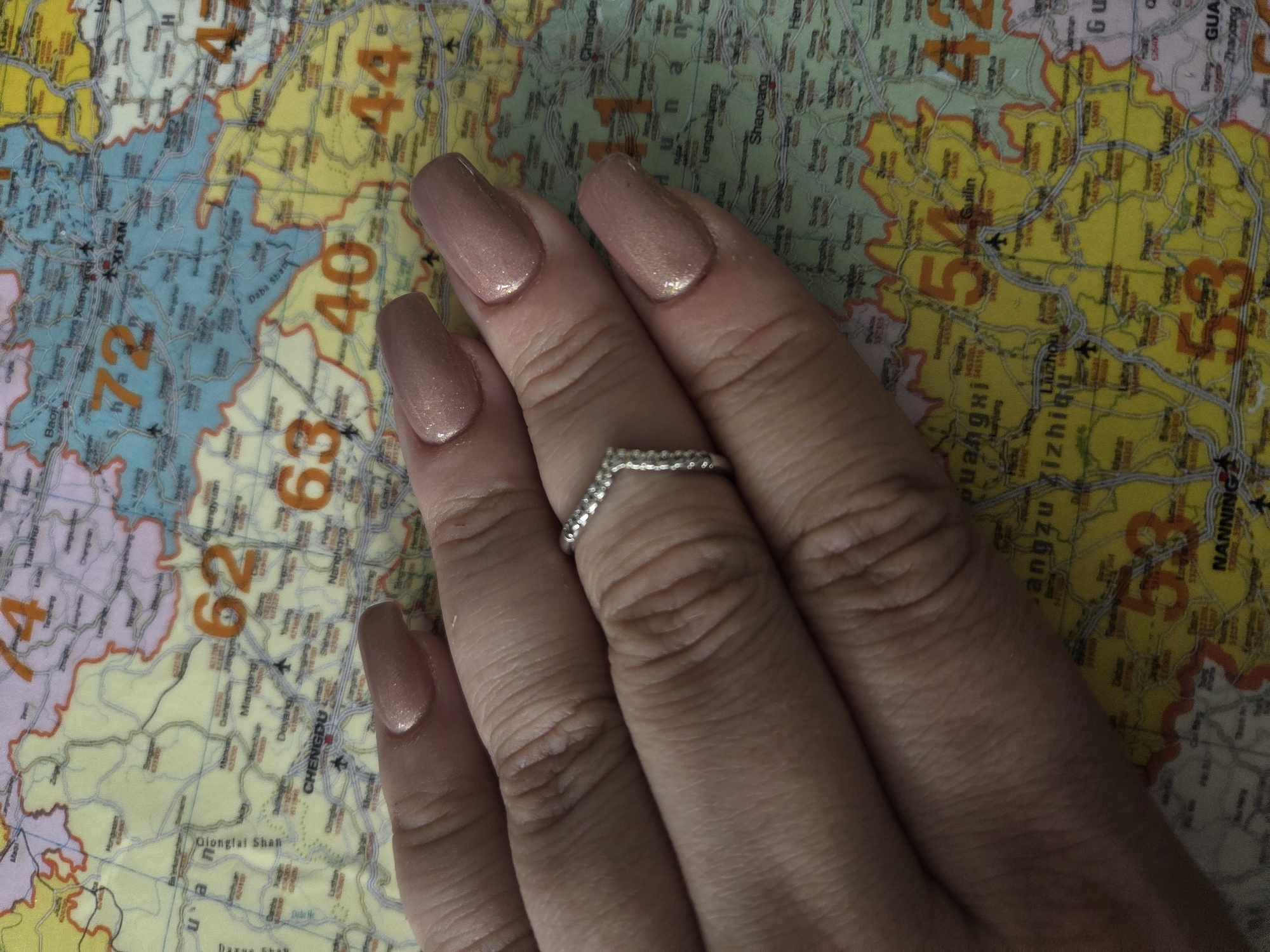 A human hand wearing a silver V-shaped ring is positioned on top of a map. The fingernails are painted a light brown color. The map features various cities and geographical areas, along with numerical designations and key points of interest. Specific locations visible on the map include Chengdu, Nanning, Guangxi, and the numerical markers 44, 54, 62, 63, 40, 53, and 72. The surrounding environment appears to be a flat surface where the map is laid out.