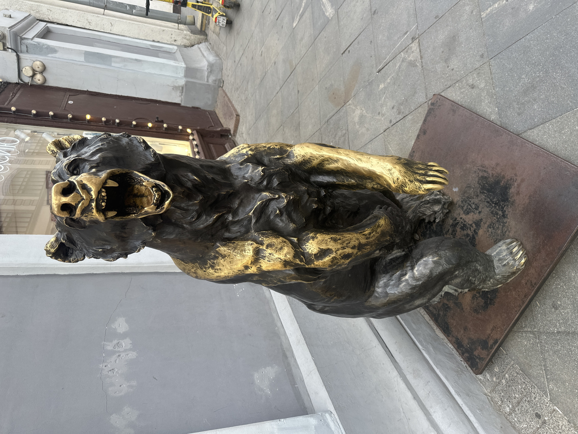 A bronze statue of a bear with its mouth open in a roar is positioned on a square metal base in front of a building. The bear is dark in color with gold accents on its face, paws, and along its musculature. The building behind the bear has large windows, a brown door, and is decorated with string lights. The ground in front of the building is paved with gray square tiles. To the right of the statue, two yellow electric scooters are parked. The scene appears to be on a street in Moscow, Russia, possibly near a commercial establishment.