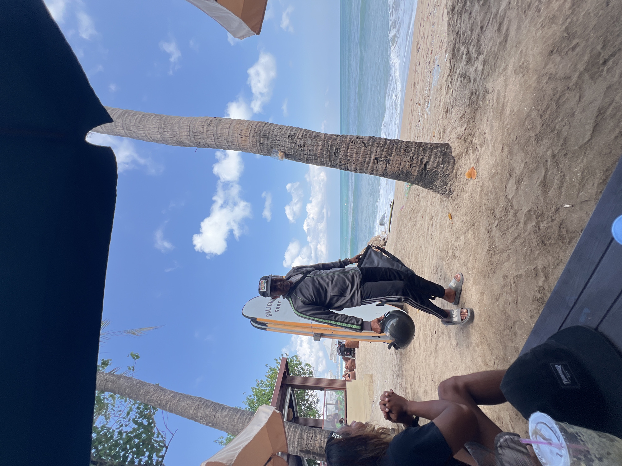 A man stands on a sandy beach in Kuta, Indonesia, near a surfboard. He is wearing a grey jacket with green stripes, black track pants, and grey sandals. He is holding a black helmet in his left hand. To his left, a person with long hair sits on the sand, looking towards him. Behind them, palm trees and beach umbrellas are visible. The ocean stretches out in the background under a blue sky with white clouds. A table with a drink and a hat is in the foreground. The scene appears to be a casual outdoor setting, possibly a beachside cafe or restaurant.