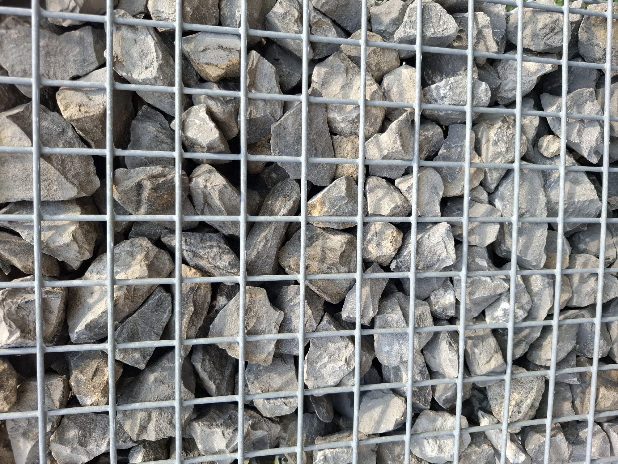 A gabion wall, filled with rough, grey stones, is captured in a close-up, full-frame shot. The stones vary in size and shape, with some exhibiting lighter grey or beige tones due to surface variations or light reflection. The gabion is constructed from a grid of thin, silver-colored metal bars, forming rectangular openings through which the stones are visible. The lighting suggests an outdoor environment, with shadows indicating a bright, possibly sunny, day. The image appears to be taken in Eschenbach (LU), Switzerland, likely in a public space where gabion walls are commonly used for landscaping or retaining earth. There are no people or animals present in the frame.