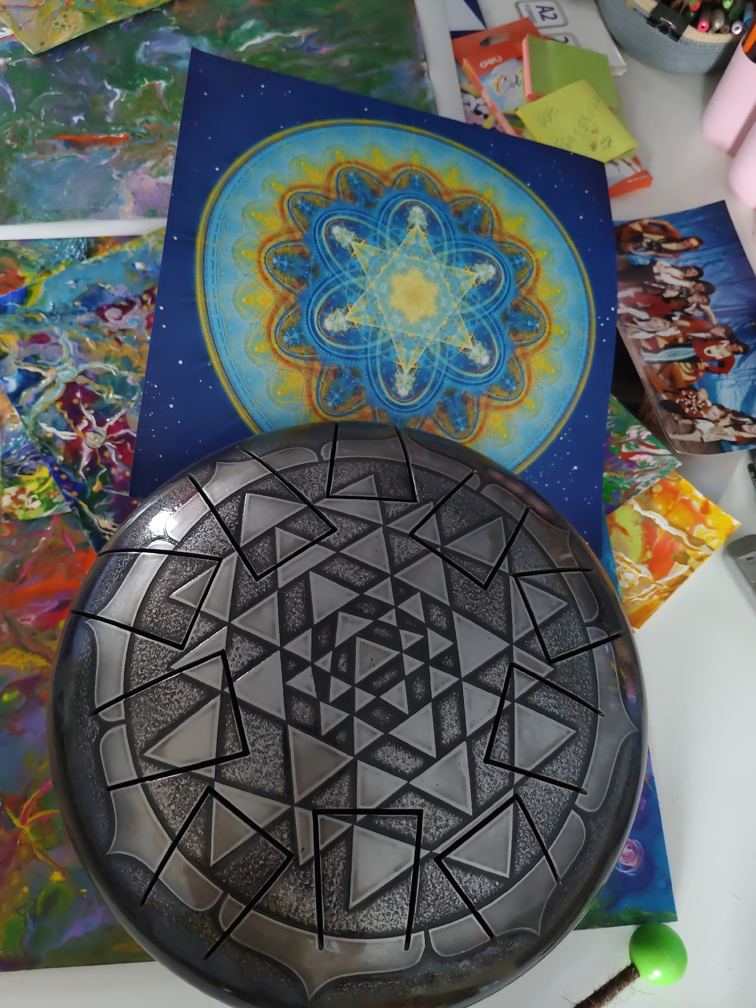 The image shows a close-up, top-down view of a steel tongue drum with a complex geometric pattern on its surface, set against a background of various artistic prints and stationery items. The drum is dark gray with lighter gray and black etched patterns that form a tessellating design of triangles and squares. Behind the drum, a large blue print with a vibrant mandala design in yellow, blue, and orange is visible. To the right of the blue print, a smaller print depicting several figures in an artistic style is present. Scattered around these prints are various stationery items, including colorful sticky notes and what appear to be markers in a light blue basket. In the lower right corner, a bright green spherical object on a brown stick is partially visible. The overall environment suggests an artist's workspace or a creative setting. There is no identifiable location in Rostov-on-Don, Russia, depicted in this image.