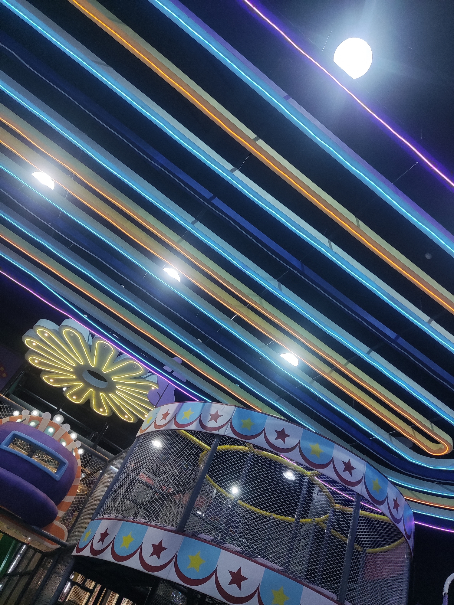 The image shows a colorful interior with multiple neon lights in shades of blue, purple, and orange running diagonally across the ceiling. In the foreground, there is a section of a children's play area, possibly an indoor playground. This section features a netted enclosure with a decorative border featuring red stars and yellow stars on a light blue background, reminiscent of a circus tent. To the left, a portion of a purple structure with a rectangular opening is visible, also part of the play area. Above the play structure, a large, yellow, flower-shaped neon light is illuminated. The lighting suggests this is an indoor recreational facility, likely a family entertainment center or a themed play space. The overall impression is one of a vibrant and playful atmosphere. The location is identified as a children's play area in Sidoarjo, Indonesia.