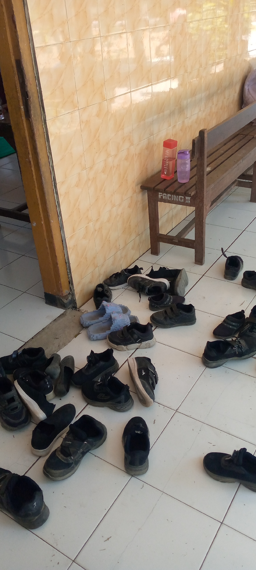 The image depicts an outdoor scene in Pacing, Indonesia, likely near a building with tiled walls and a wooden bench. A collection of black shoes and blue sandals are scattered on the white tiled floor. Some of the shoes appear to be athletic shoes, while others are more casual. Two water bottles, one red and one purple, are placed on the bench, with the word "MY BOTTLE" visible on the red one. The word "PACING" is stenciled on the front of the bench. The environment appears to be a communal area where people might remove their shoes before entering a building.