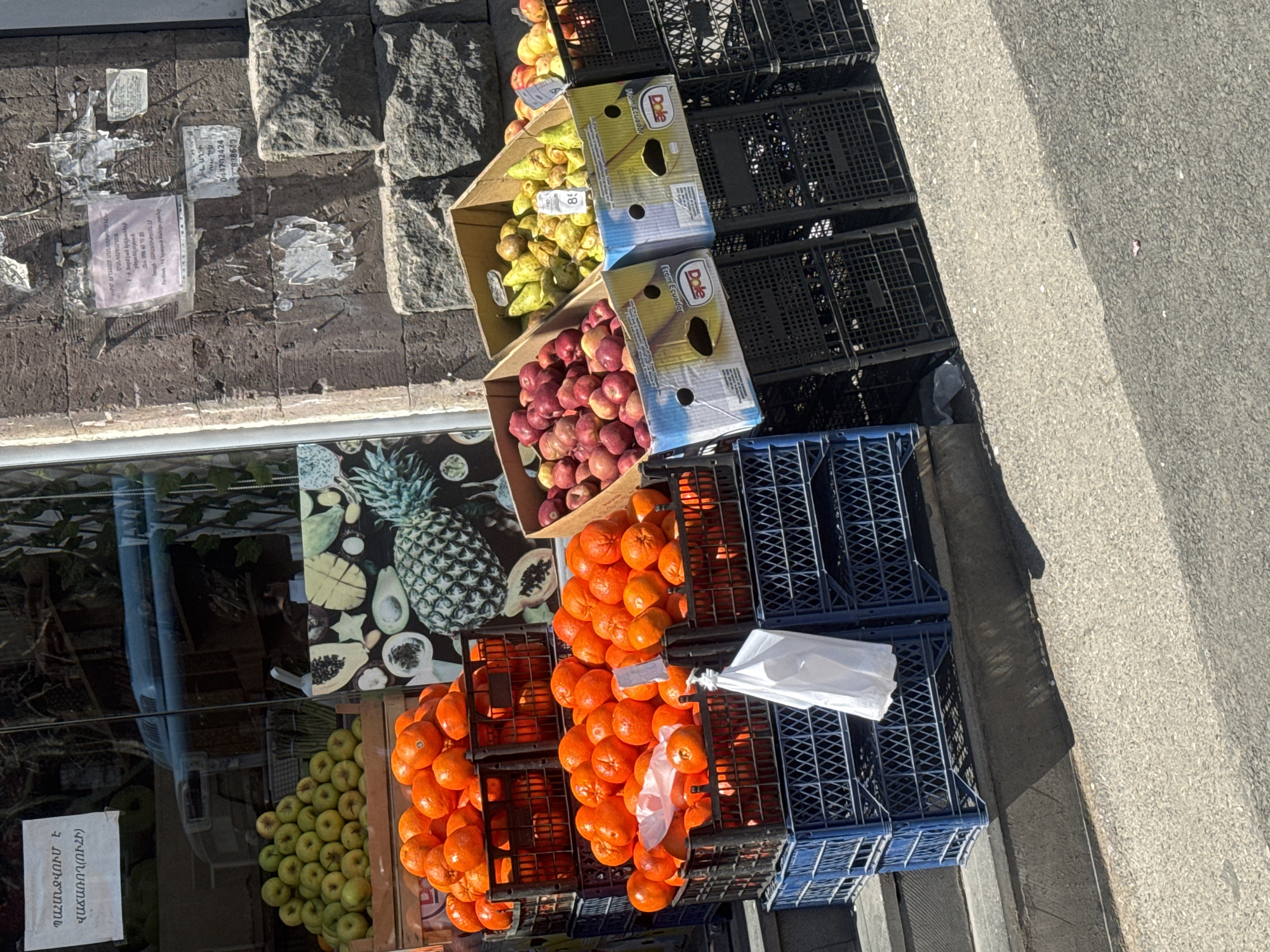 The image shows a fruit stand set up on the side of a street in Yerevan, Armenia. A variety of fruits, including apples, pears, and tangerines, are displayed in crates. The crates are stacked on top of each other. The fruit stand is located next to a building with a stone facade and peeling notices and advertisements. The sidewalk and street are visible. The image includes words in Armenian, implying the location. The brand Dole is also visible on the boxes.
