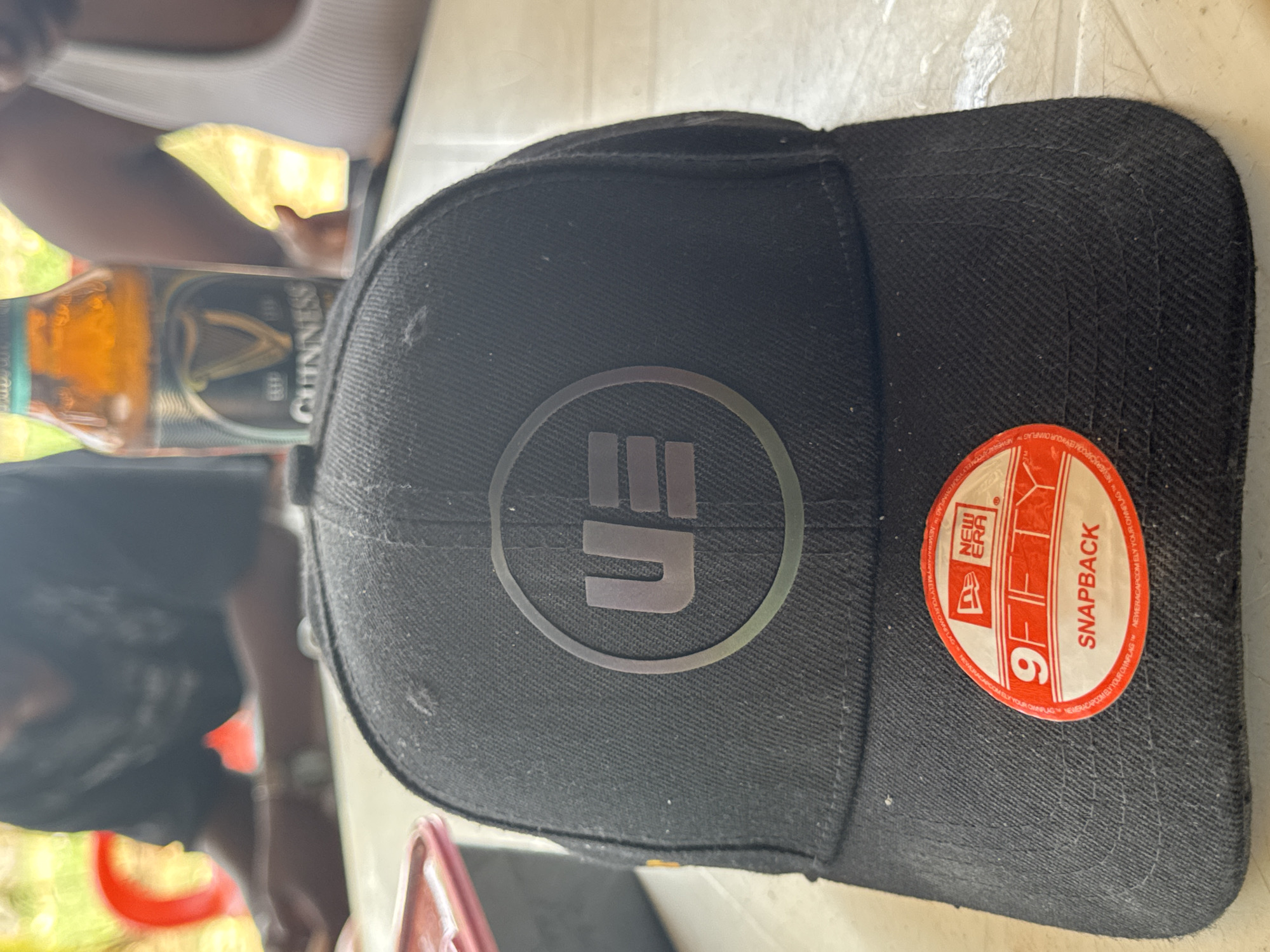 A black baseball cap, viewed from an overhead perspective, is the central subject of the image. The cap rests on a light-colored, possibly plastic, table surface. On the front of the cap, there is a circular, iridescent grey logo featuring stylized letters that resemble "E" and "Π". A prominent red New Era sticker is affixed to the right side of the cap's brim, clearly displaying "NEW ERA", "9FIFTY", and "SNAPBACK" in white text, along with smaller, repeated text including "NEWERACAPCOM" and "YOUR OWNFLAG".
In the blurred background to the left of the cap, a brown glass bottle of Guinness is partially visible, with the brand name "GUINNESS" legible on its label. Further in the background, blurred forms of people are discernible, along with hints of green and yellow, suggesting outdoor foliage. This indicates an outdoor or semi-outdoor setting, possibly a casual gathering during daytime in Kurunduma, Nigeria. Other blurred objects include a yellow item near the Guinness bottle and a reddish-brown item on the table closer to the viewer.