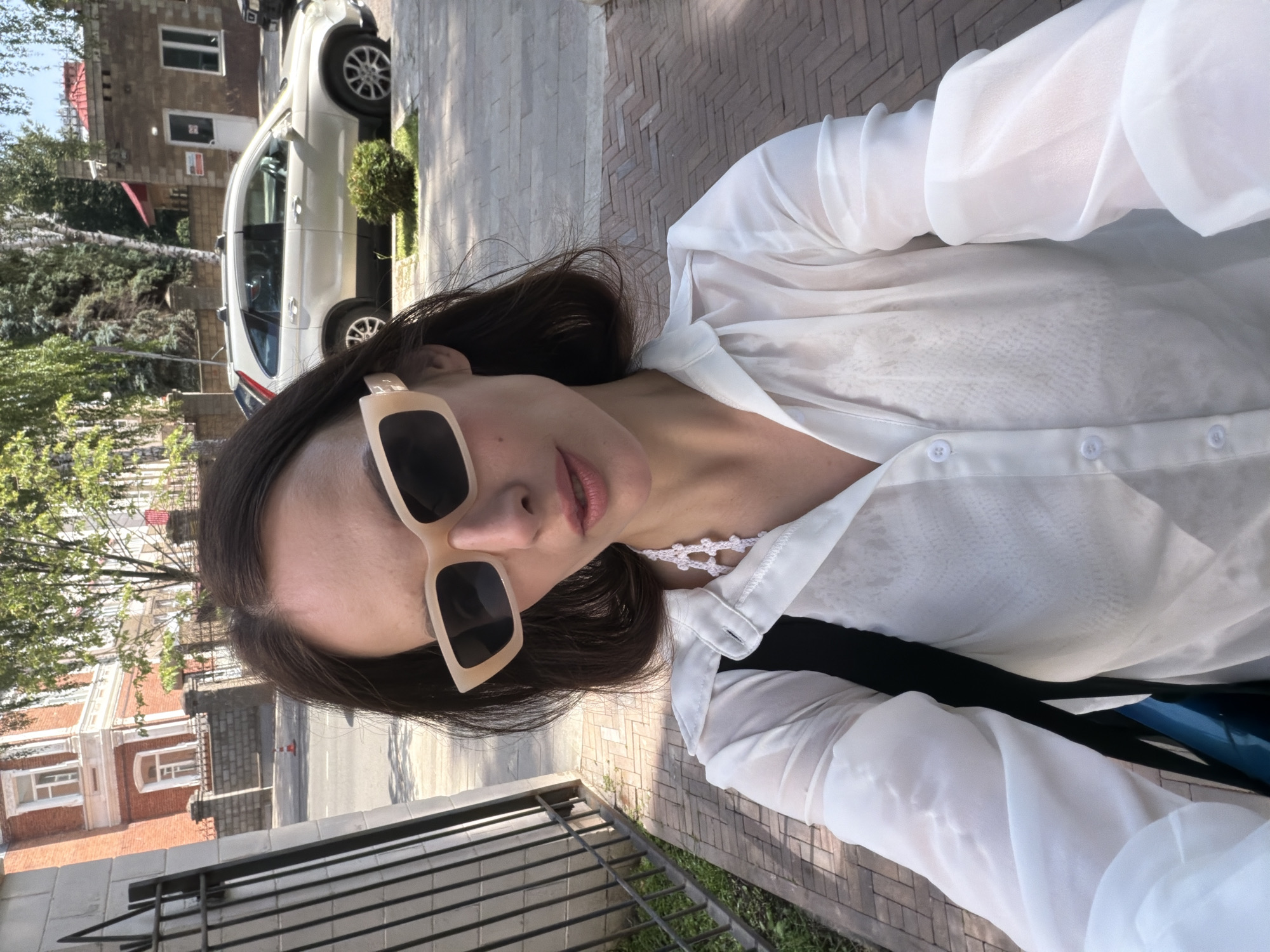 In a street scene in Ufa, Russia, a woman with dark hair and sunglasses takes a selfie. She is wearing a white, sheer blouse over a dark top, and a white crochet necklace. She has a black bag slung over her shoulder. Behind her, a white SUV is parked on the street. The background includes a brick building, a metal fence, trees, and a paved sidewalk. The weather appears to be sunny.