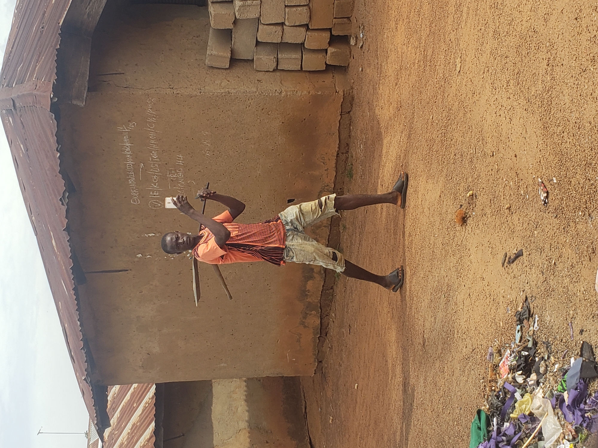 In Kachia, Nigeria, a young Black male stands in front of a mud-brick building with a corrugated metal roof. He wears an orange t-shirt with a patterned design, ripped denim shorts, and dark sandals. The man is holding a thin, dark wooden pole across his shoulders with both hands. He is looking at a white smartphone he is holding in his right hand, which is positioned in front of him. The building behind him has chalked writing on its wall. To the right of the building, a stack of gray concrete blocks is visible. The ground is composed of dirt and gravel. In the lower left corner of the image, a pile of trash, including purple fabric, plastic bags, and other debris, is present. The sky is overcast.